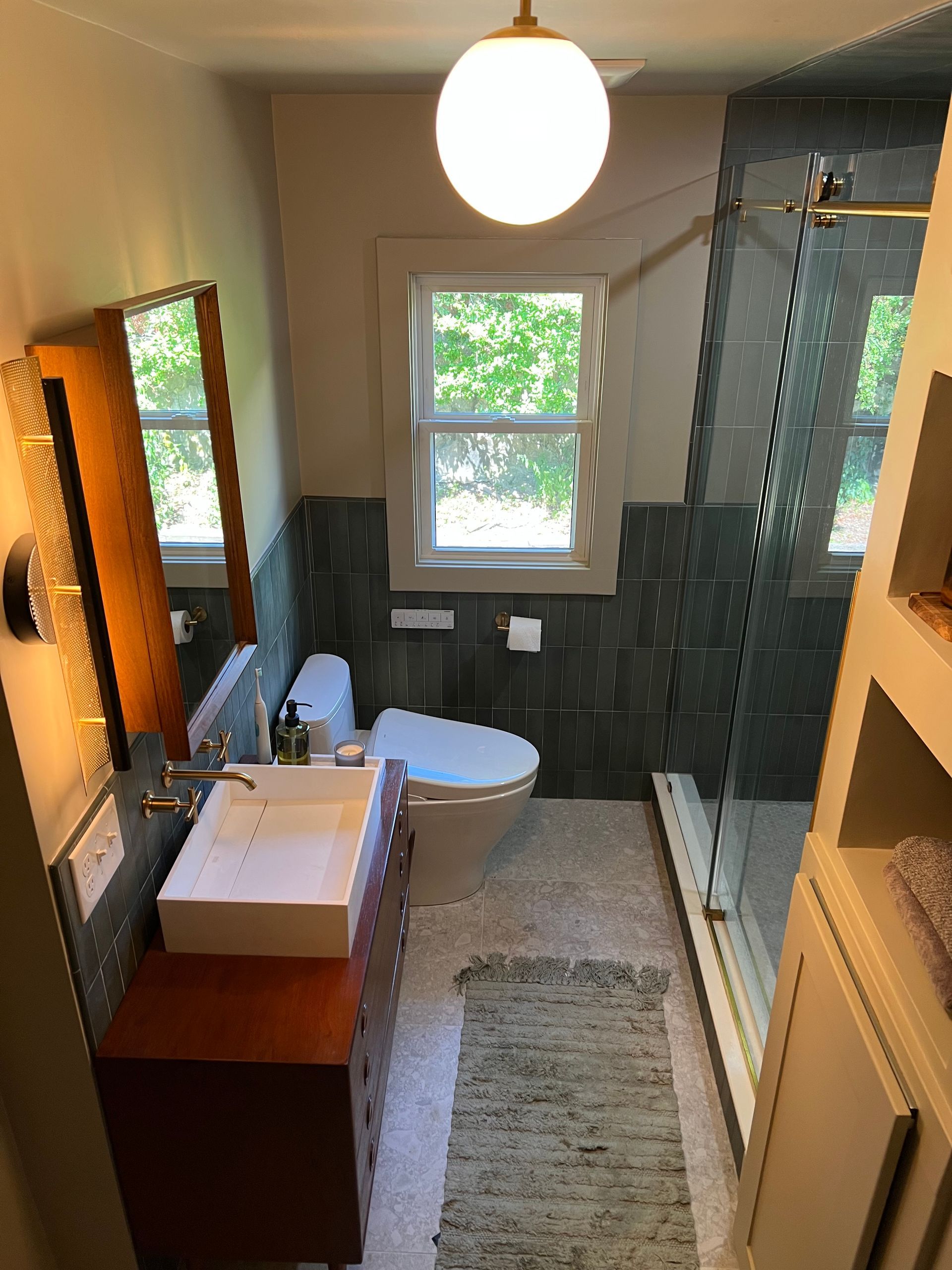 Bathroom remodel in Medford, MA showing finished modern bathroom remodel featuring a mid-century wood vanity, globe pendant light, green half-wall tile, and a glass-enclosed shower.