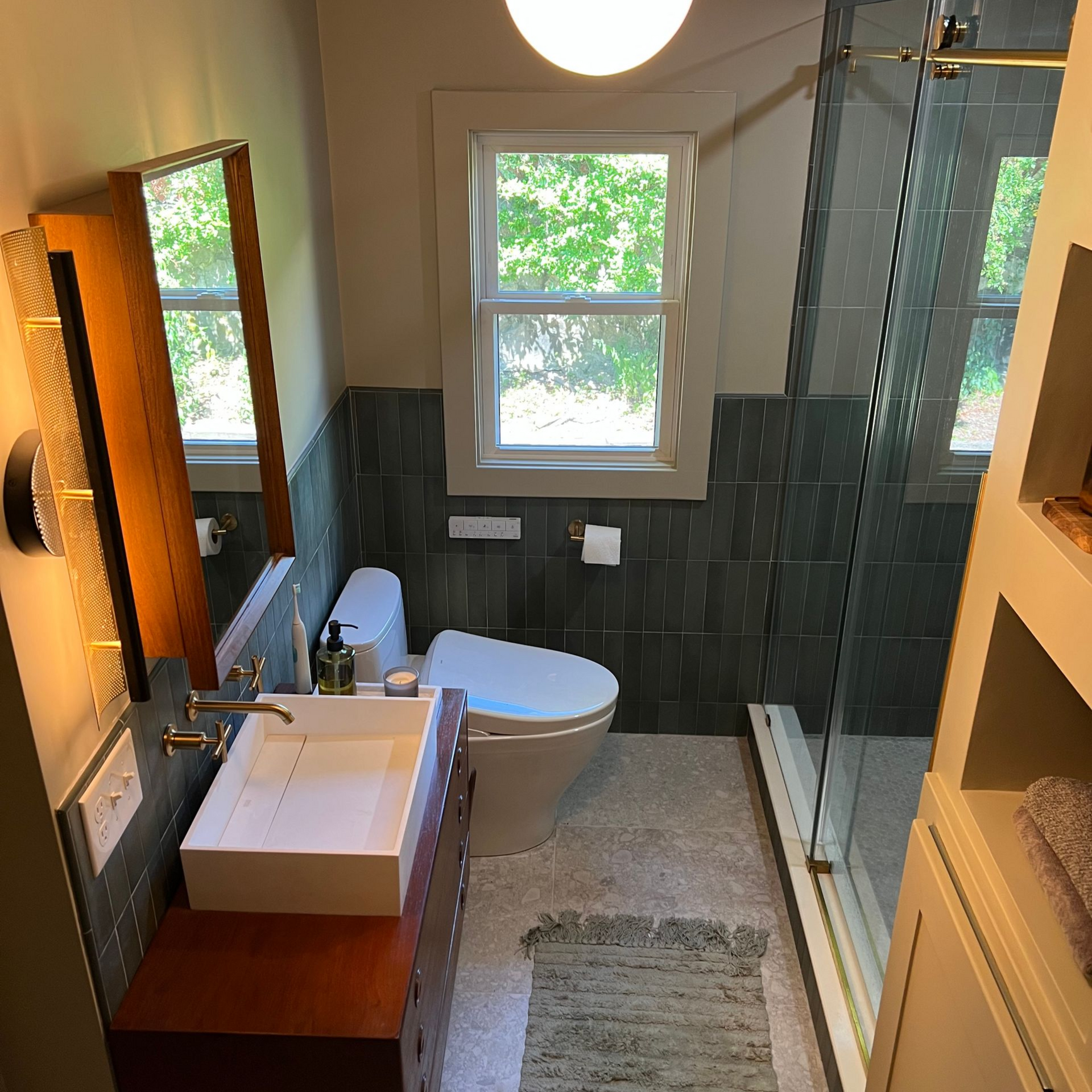 The finished modern bathroom remodel featuring a mid-century wood vanity, globe pendant light, green half-wall tile, and a glass-enclosed shower.