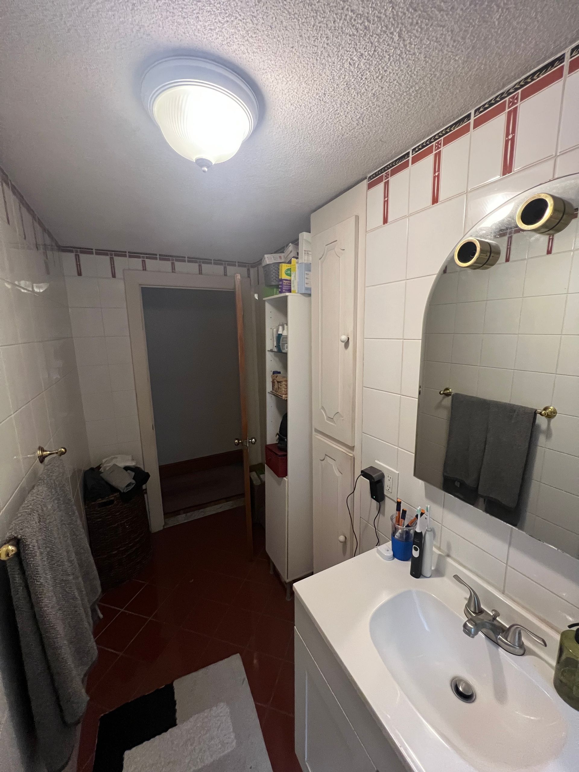 Outdated bathroom before remodel featuring white wall tiles with a red border, a basic white vanity, and red floor tiles.