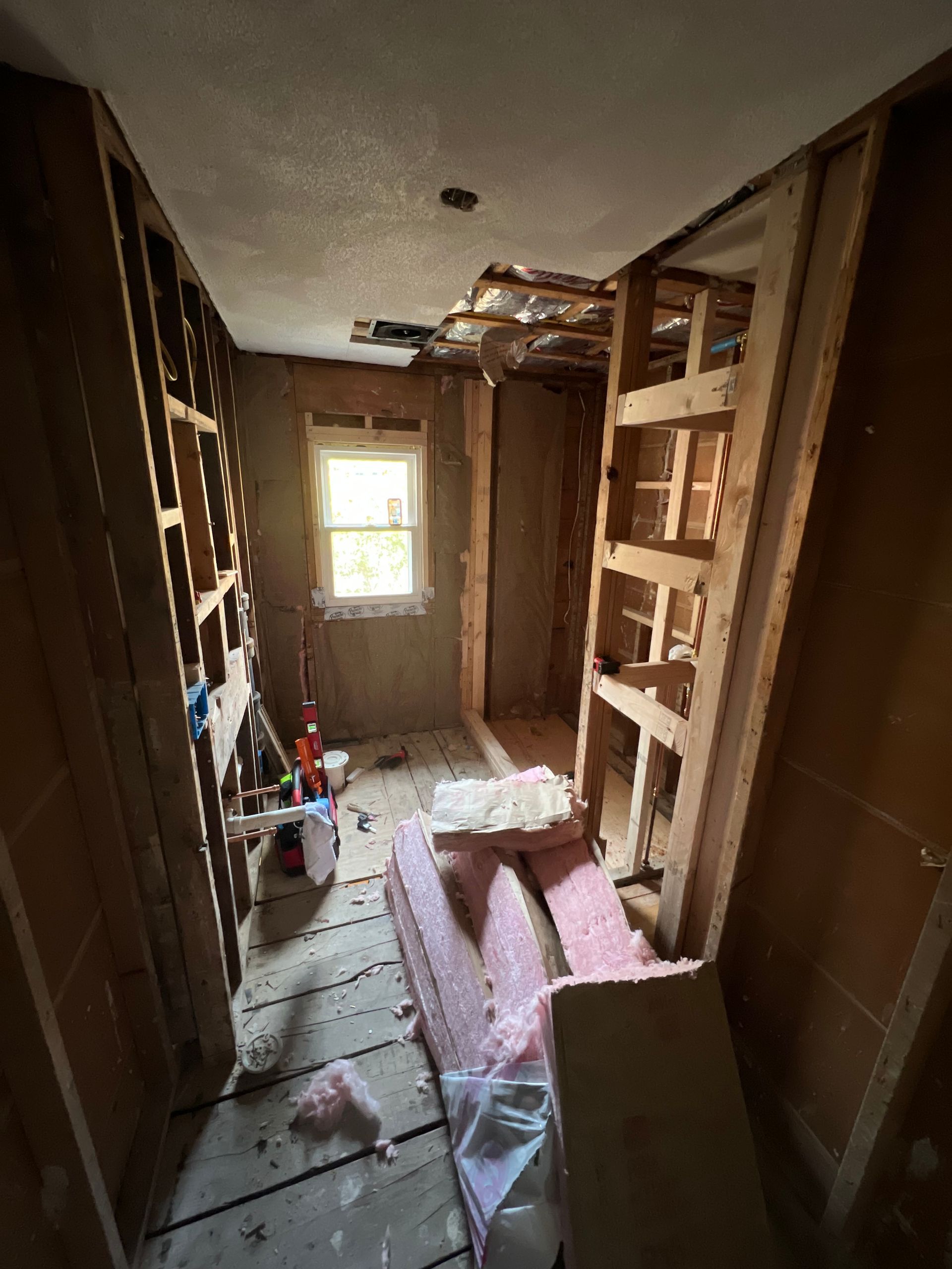Bathroom remodel demolition phase showing exposed wall studs, subfloor, and pink fiberglass insulation.