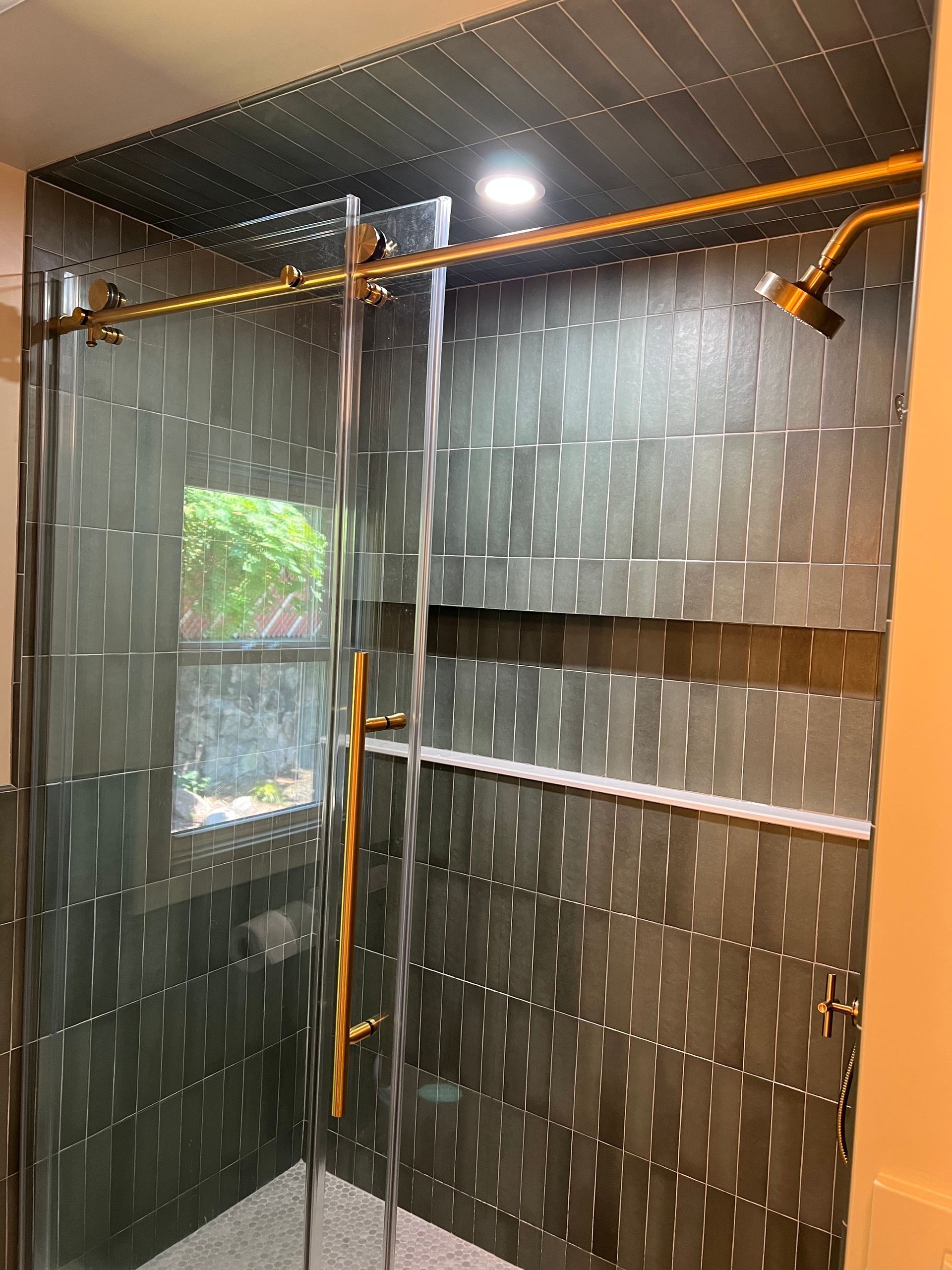 Close-up of new brushed gold rainfall showerhead and fixtures installed against dark green vertical tiles. in Medford, MA