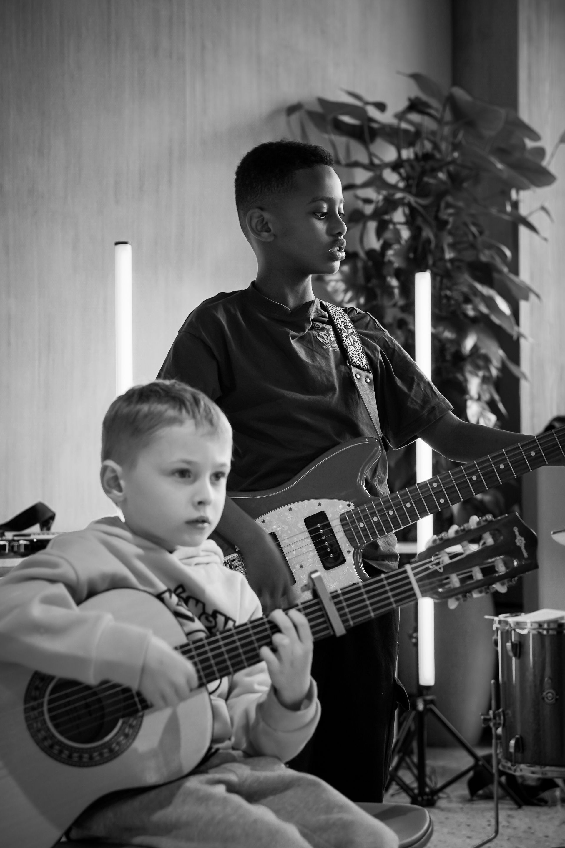 Two musicians playing guitars indoors, one seated in front and one standing behind near a plant.