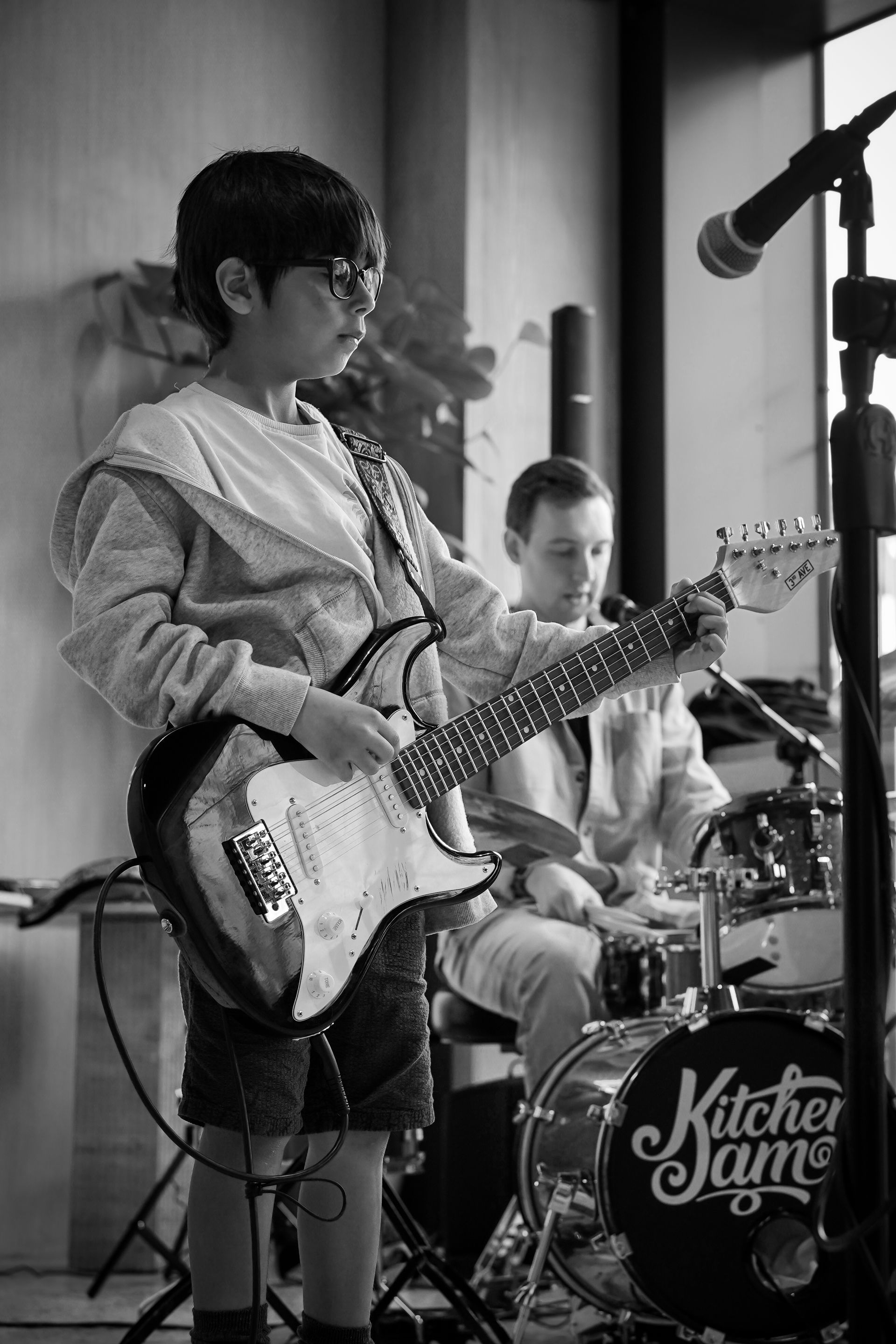 Musicians rehearsing on stage, with a guitarist in front and a drummer behind a “KitchenJam” drum kit.