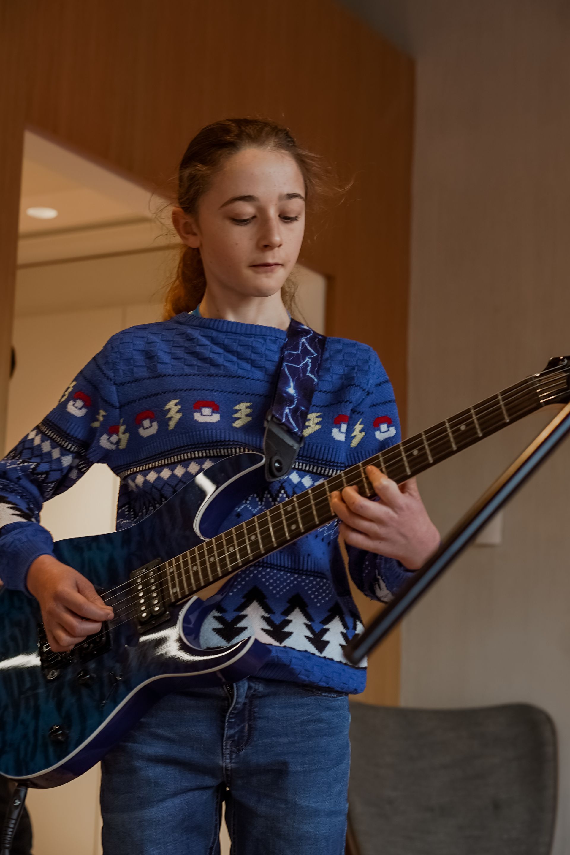 Child playing a black electric guitar in a blue sweater indoors
