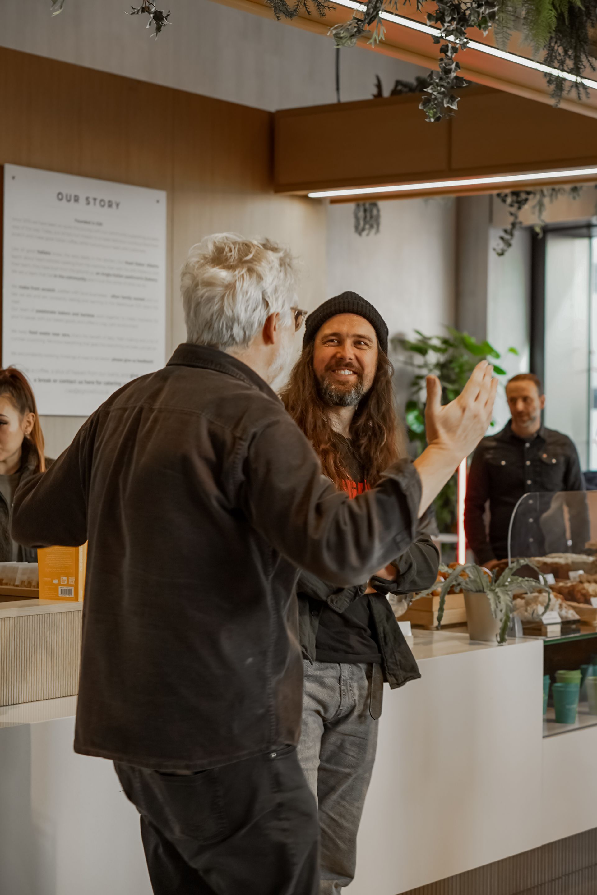 Two men chatting in a café, one gesturing with a drink near the counter.