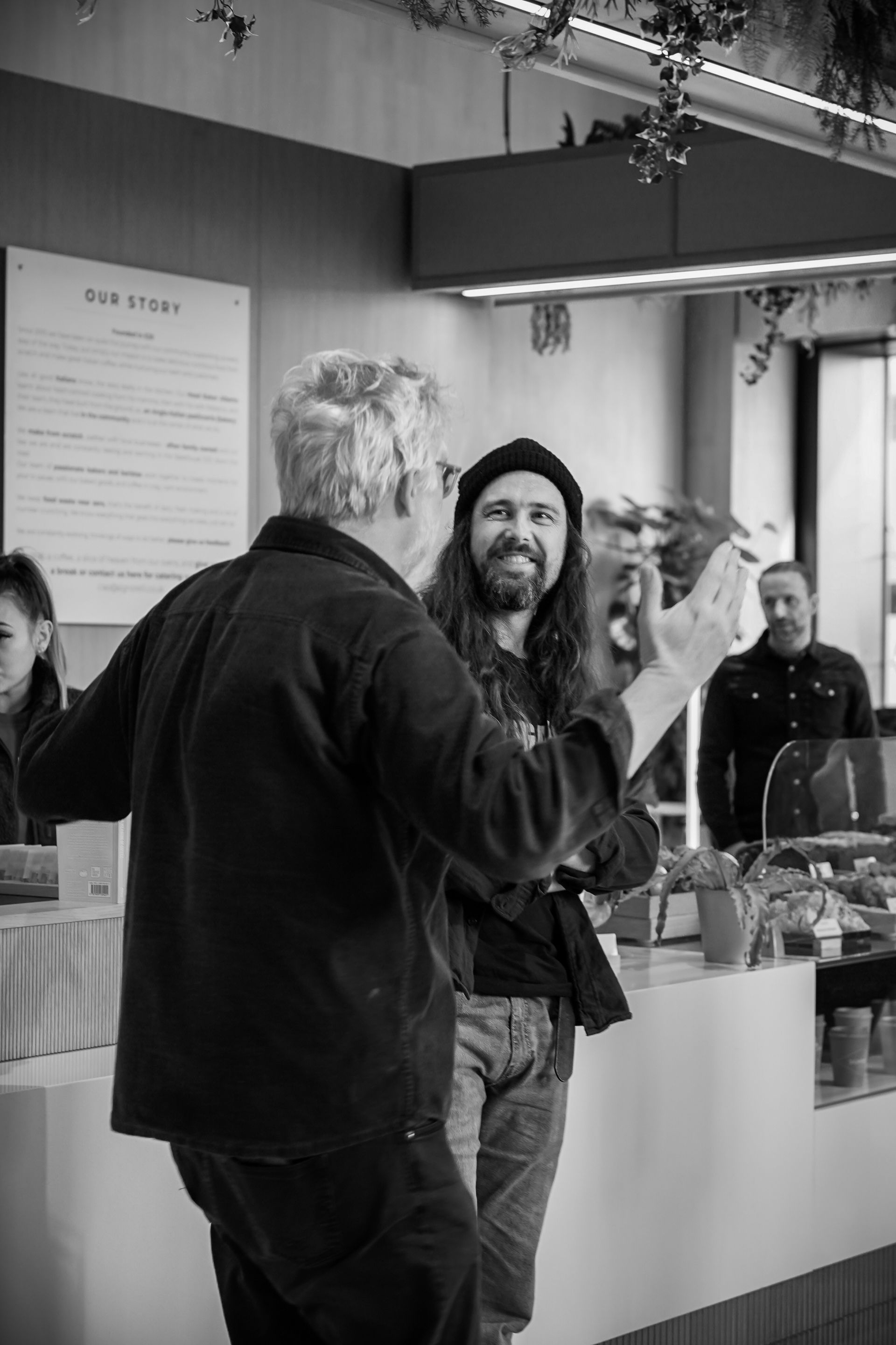 Two people talking inside a coffee shop, one smiling and gesturing at the counter.