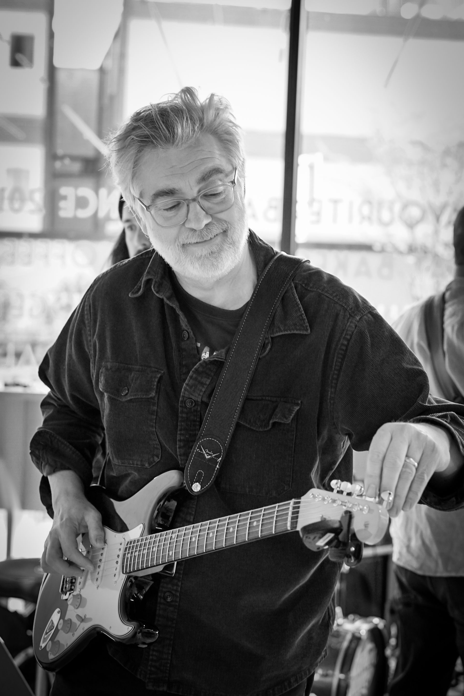 Man playing an electric guitar indoors, wearing glasses and a dark jacket.