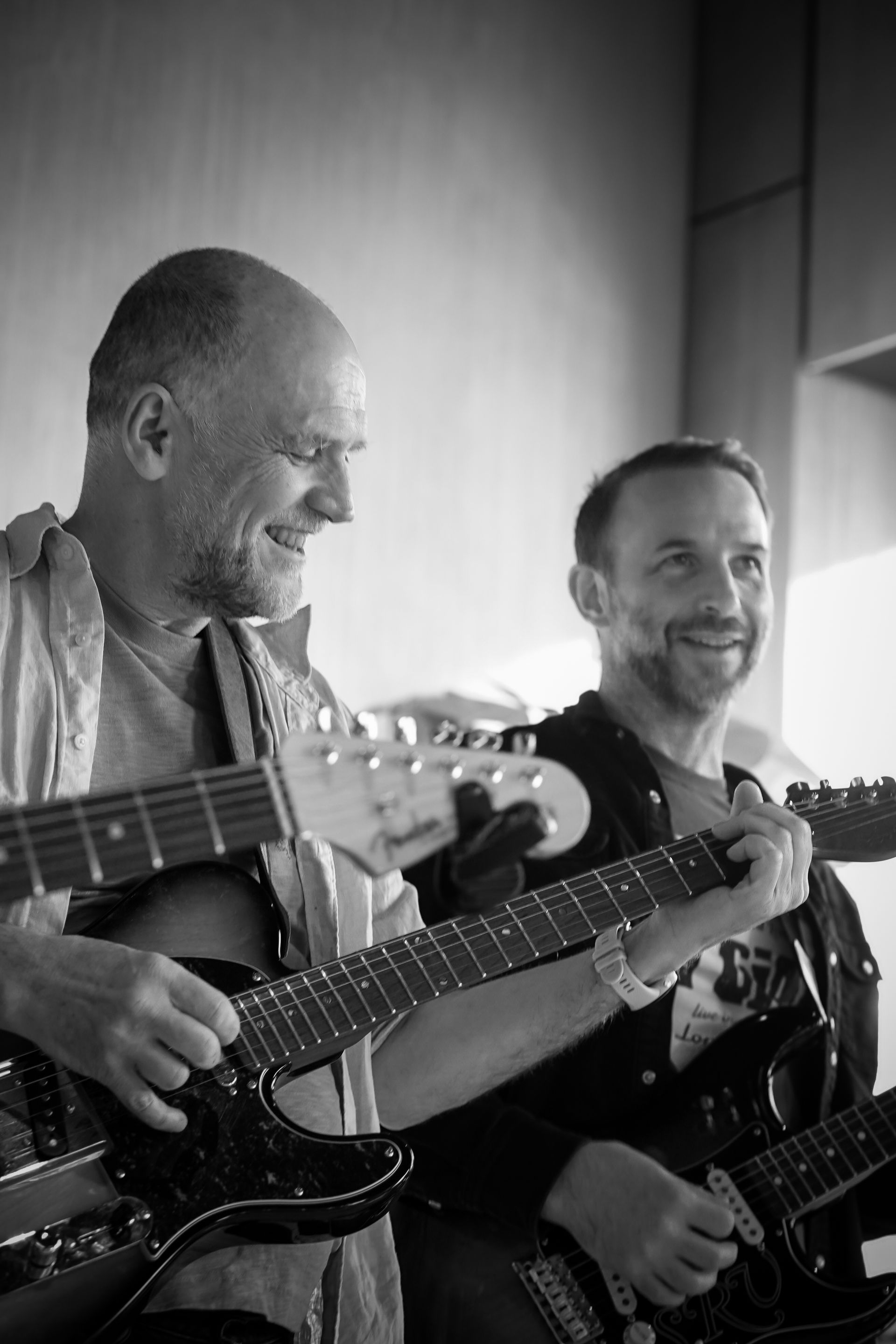 Two musicians smiling and playing electric guitars in a black-and-white indoor scene