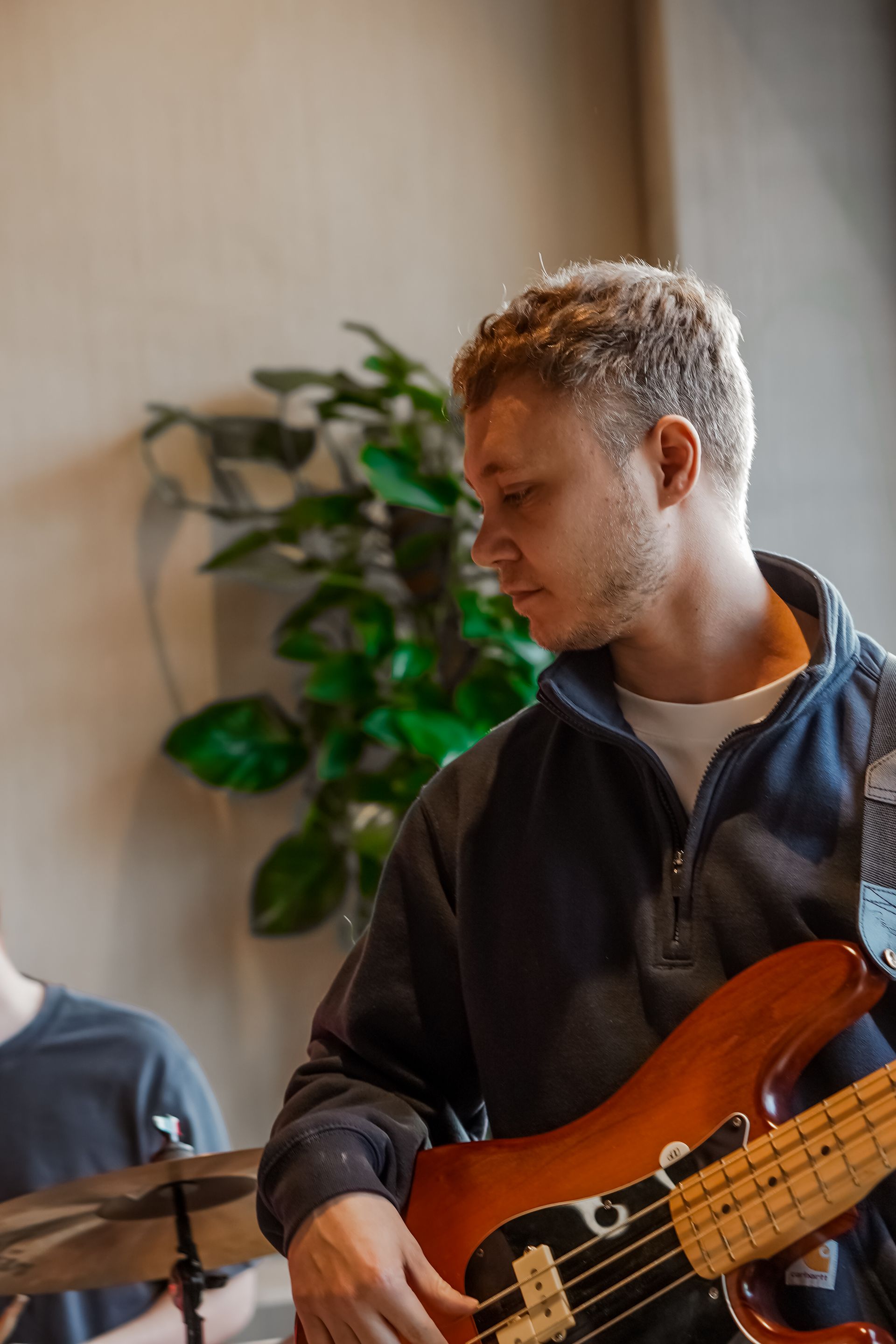 Musician playing an orange bass guitar beside a drum set indoors