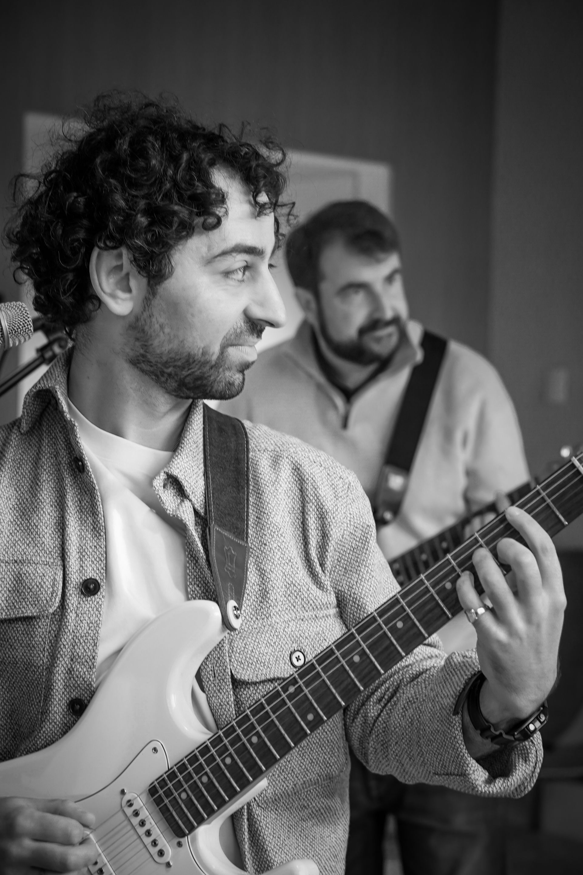Two musicians indoors, one playing electric guitar in the foreground with another blurred behind him.