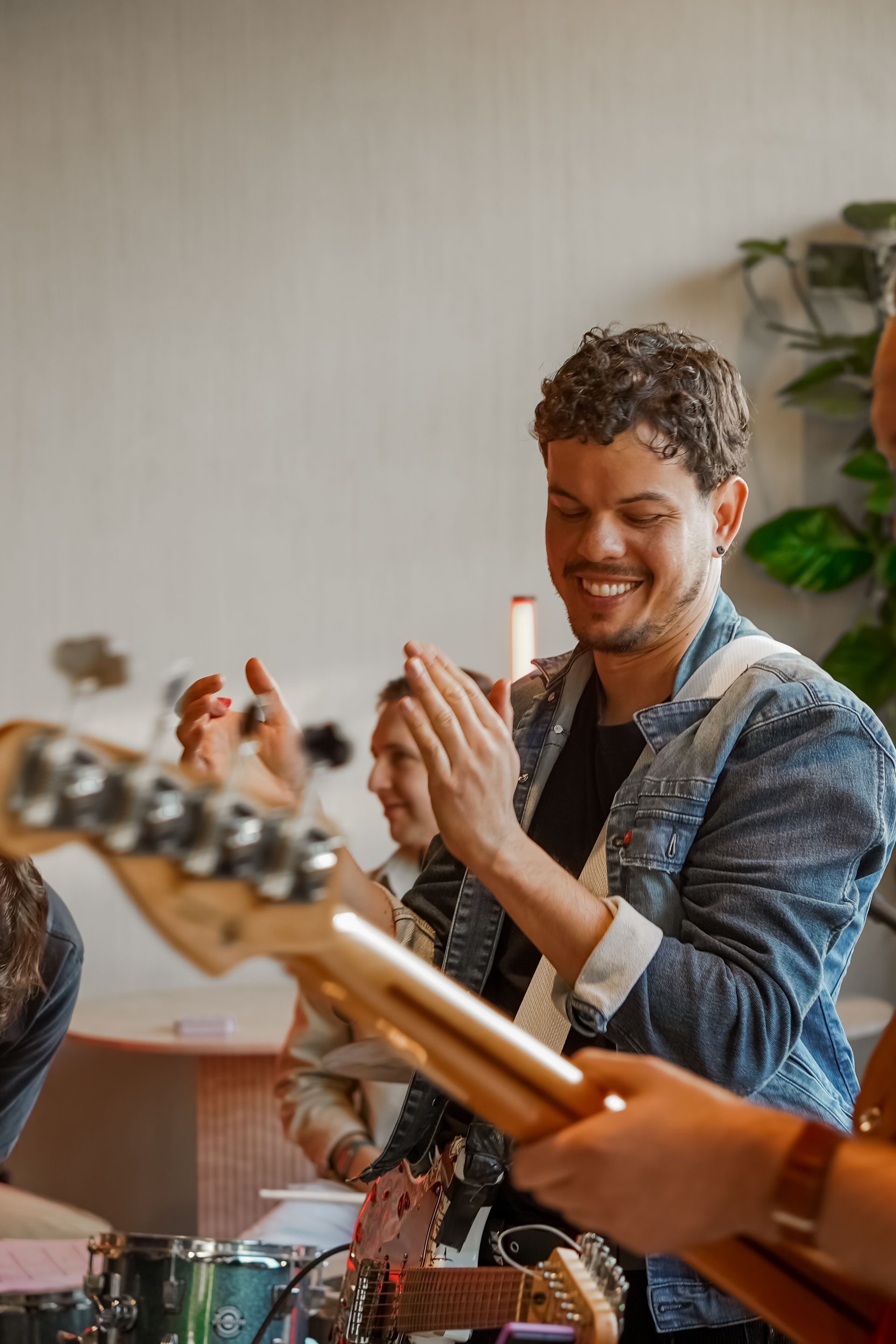 Man smiling and clapping at a music gathering, with guitars in the foreground and a plant in the background