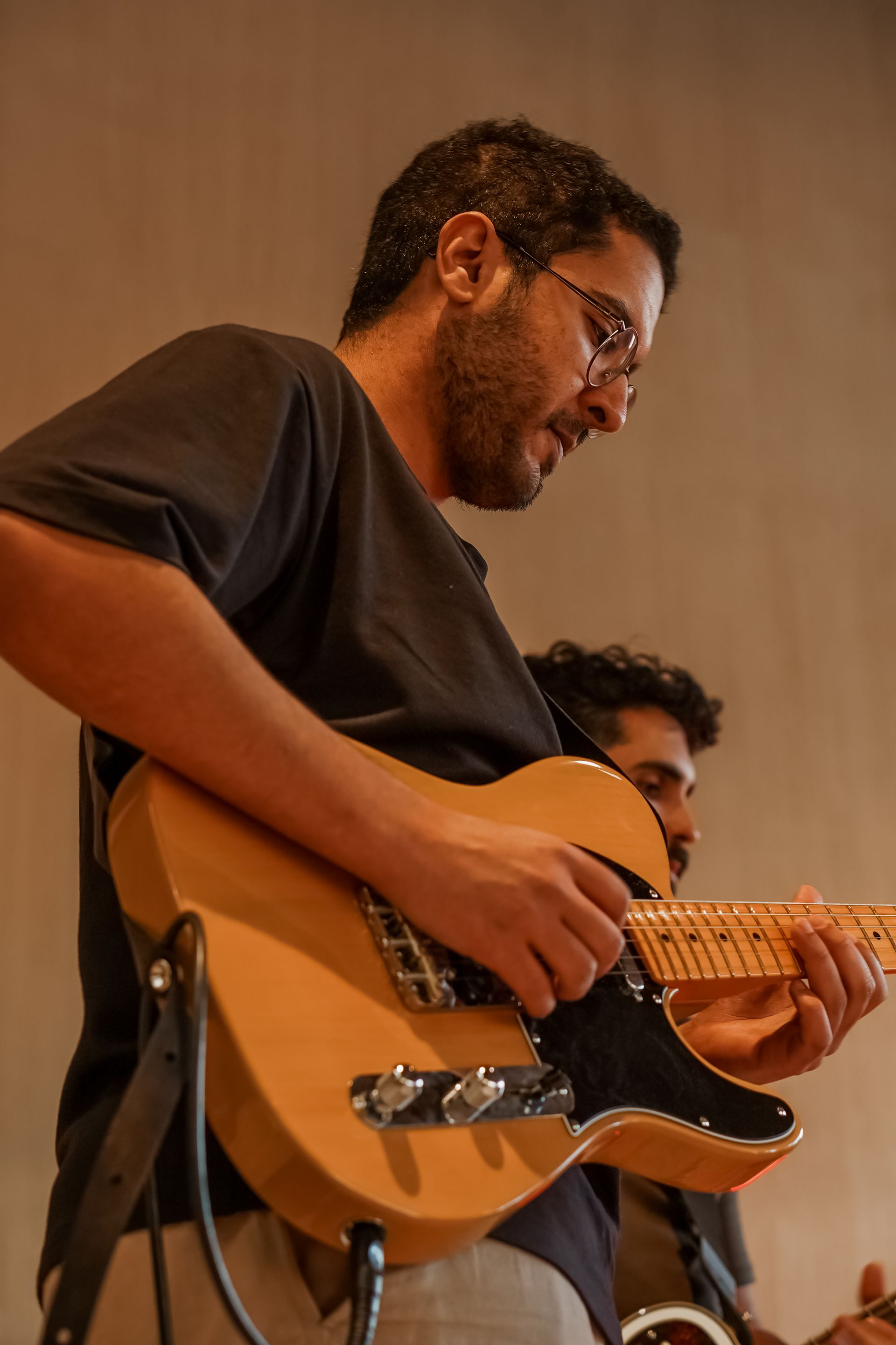 Musician playing a sunburst electric guitar on stage, with another person partially visible behind him.