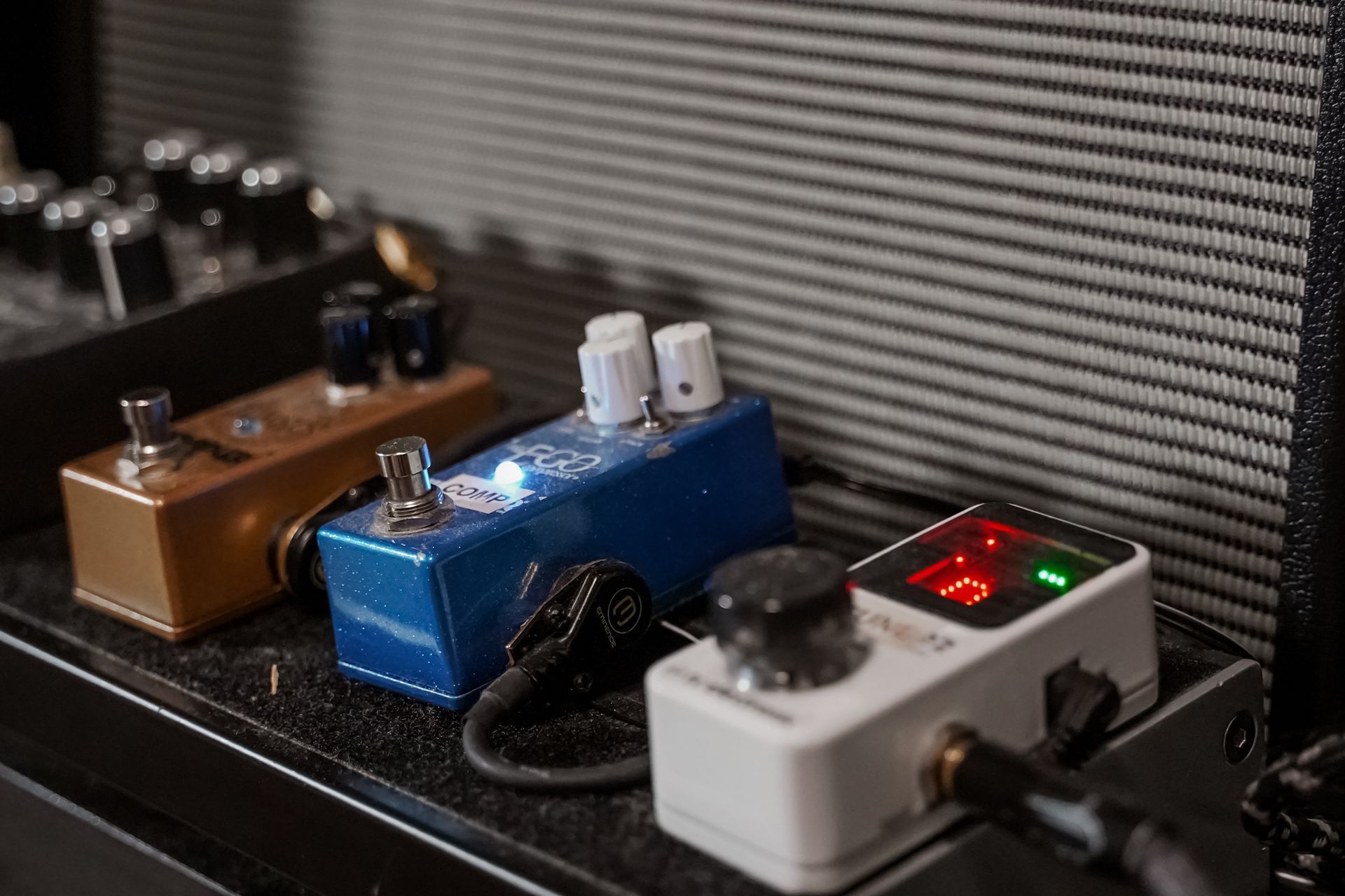 Three guitar effect pedals on a shelf, including blue, brown, and white pedals with lit red and green LEDs.