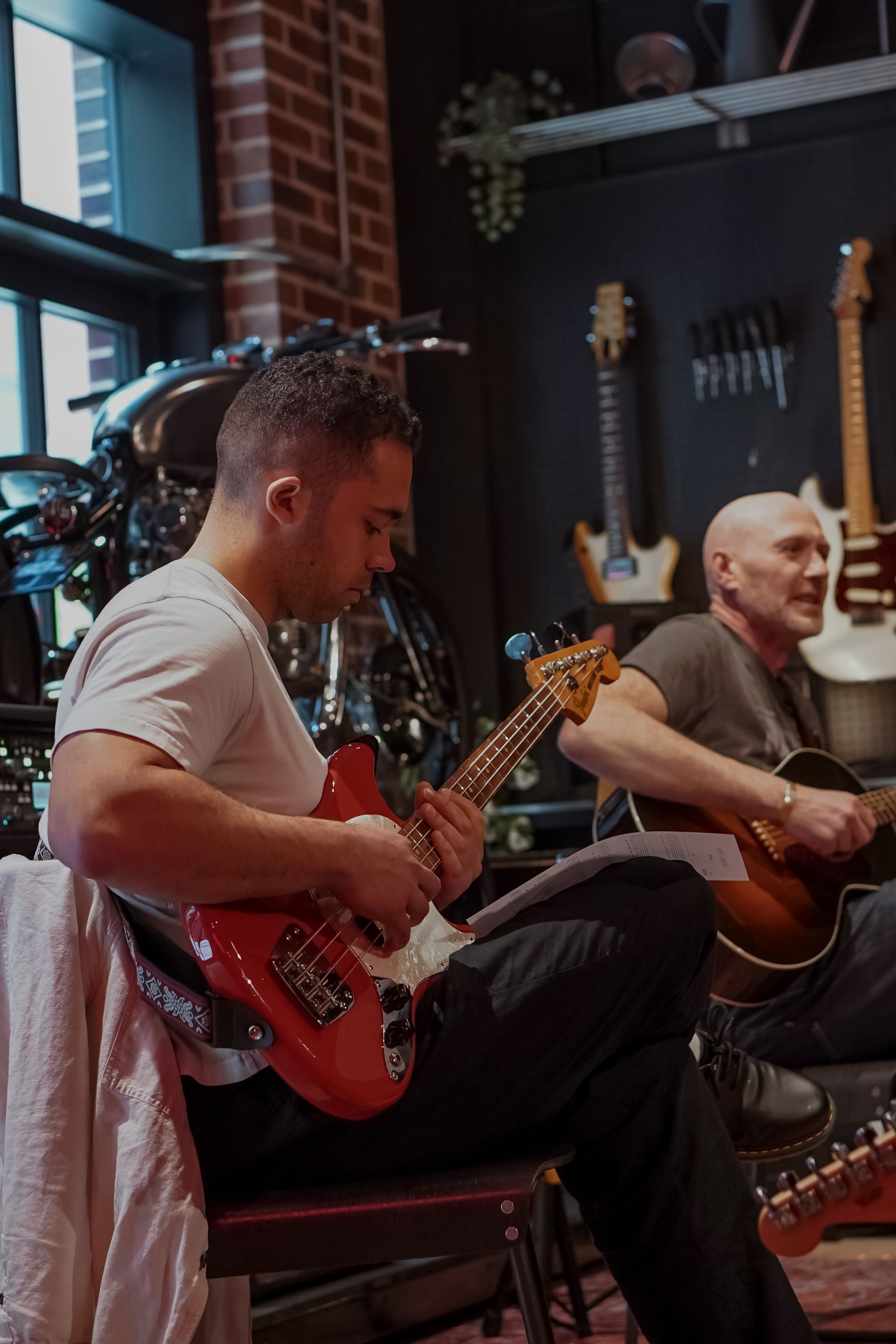 Two musicians play acoustic guitars in a cozy studio with brick walls and hanging instruments.