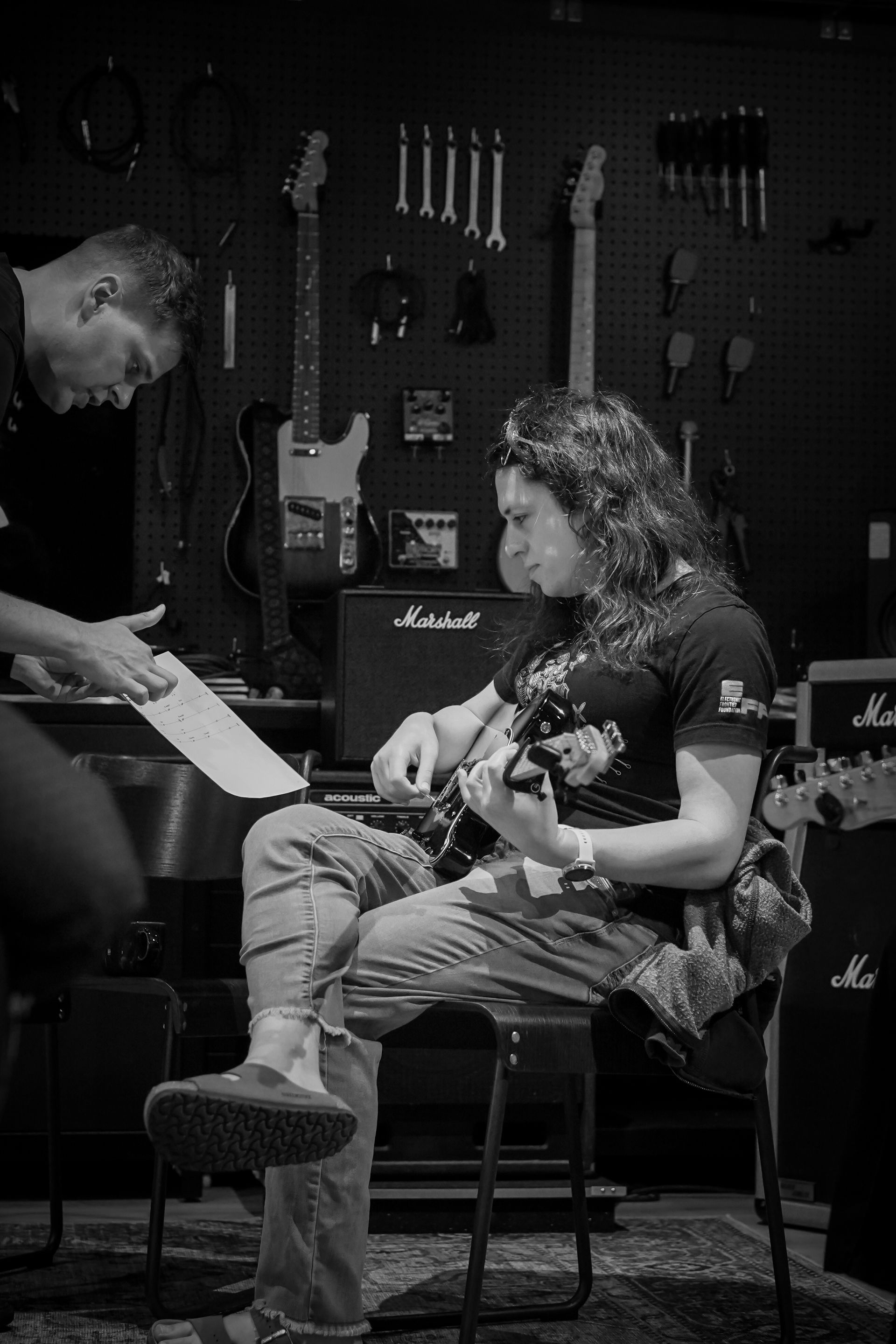 Musician playing guitar while another person leans in to speak in a guitar shop or studio