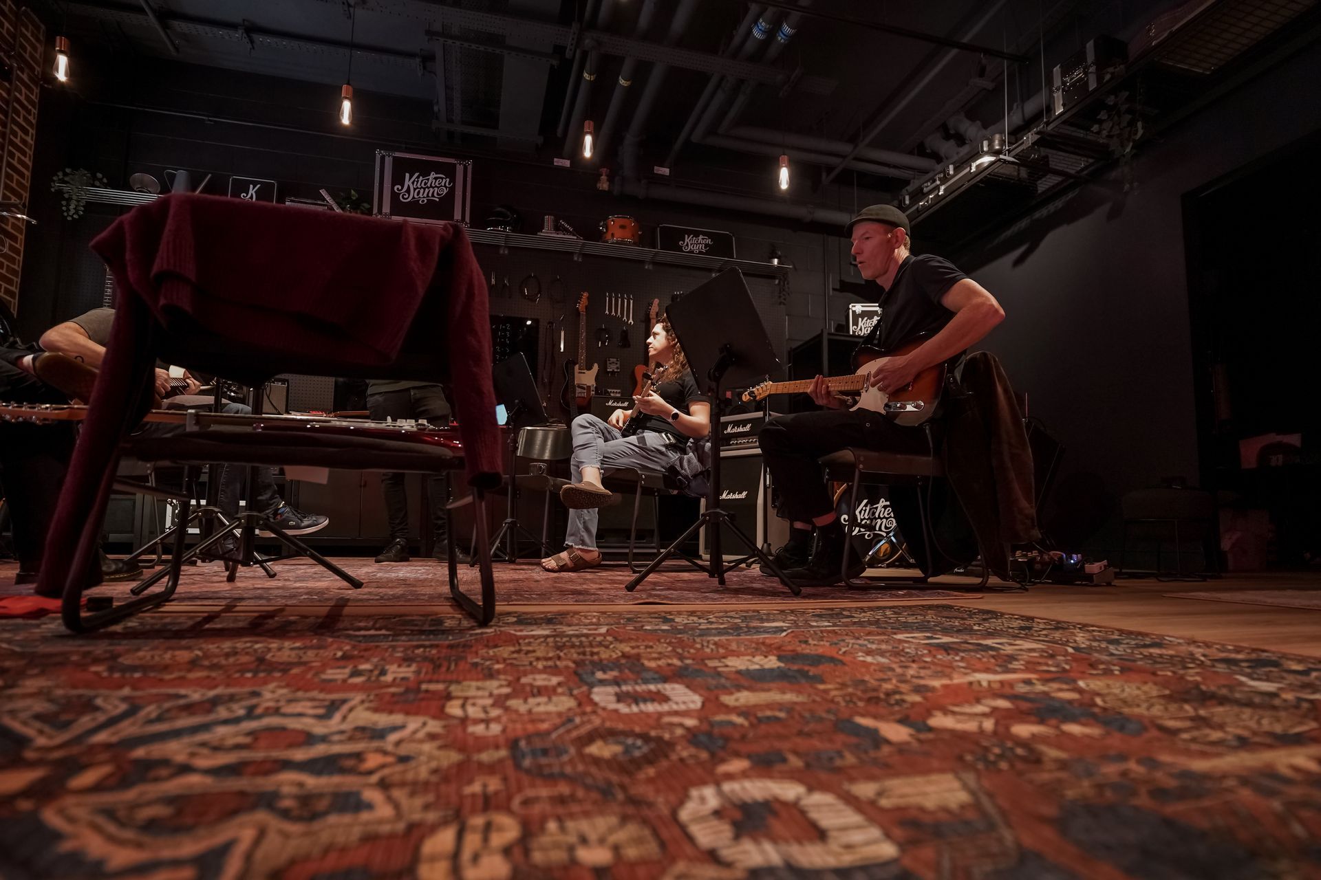 Musicians rehearsing on a dim stage with drums, chairs, and instruments on a patterned rug