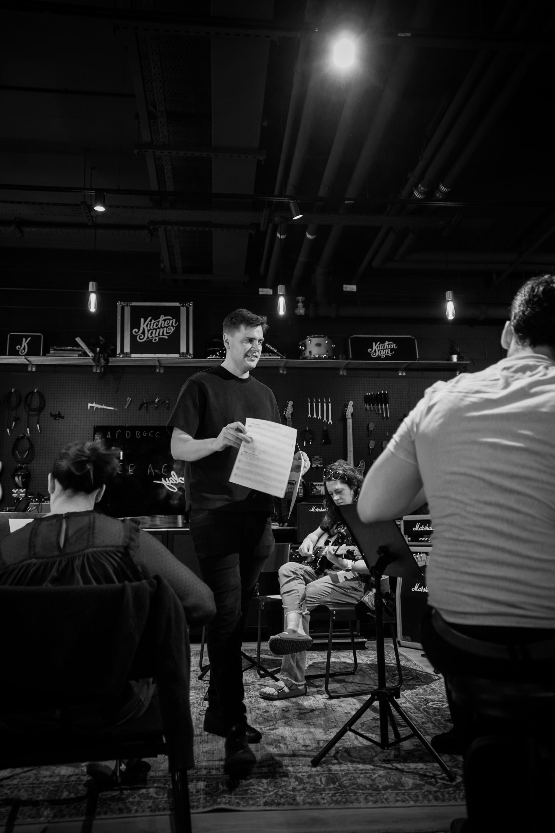 Man holding papers speaks on a stage in a dimly lit venue, with seated audience members in the foreground.