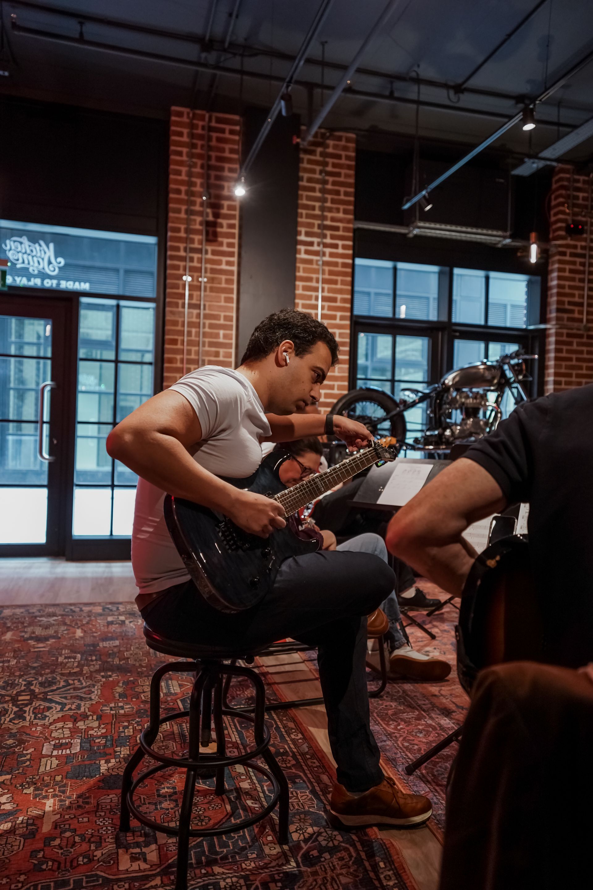 Man seated in a brick-walled studio playing a black electric guitar, with drums and windows in the background