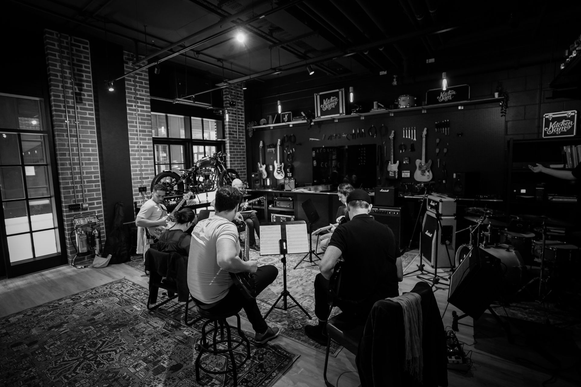 A group of musicians playing acoustic guitars while sitting in a dimly lit, brick-walled studio filled with instruments.