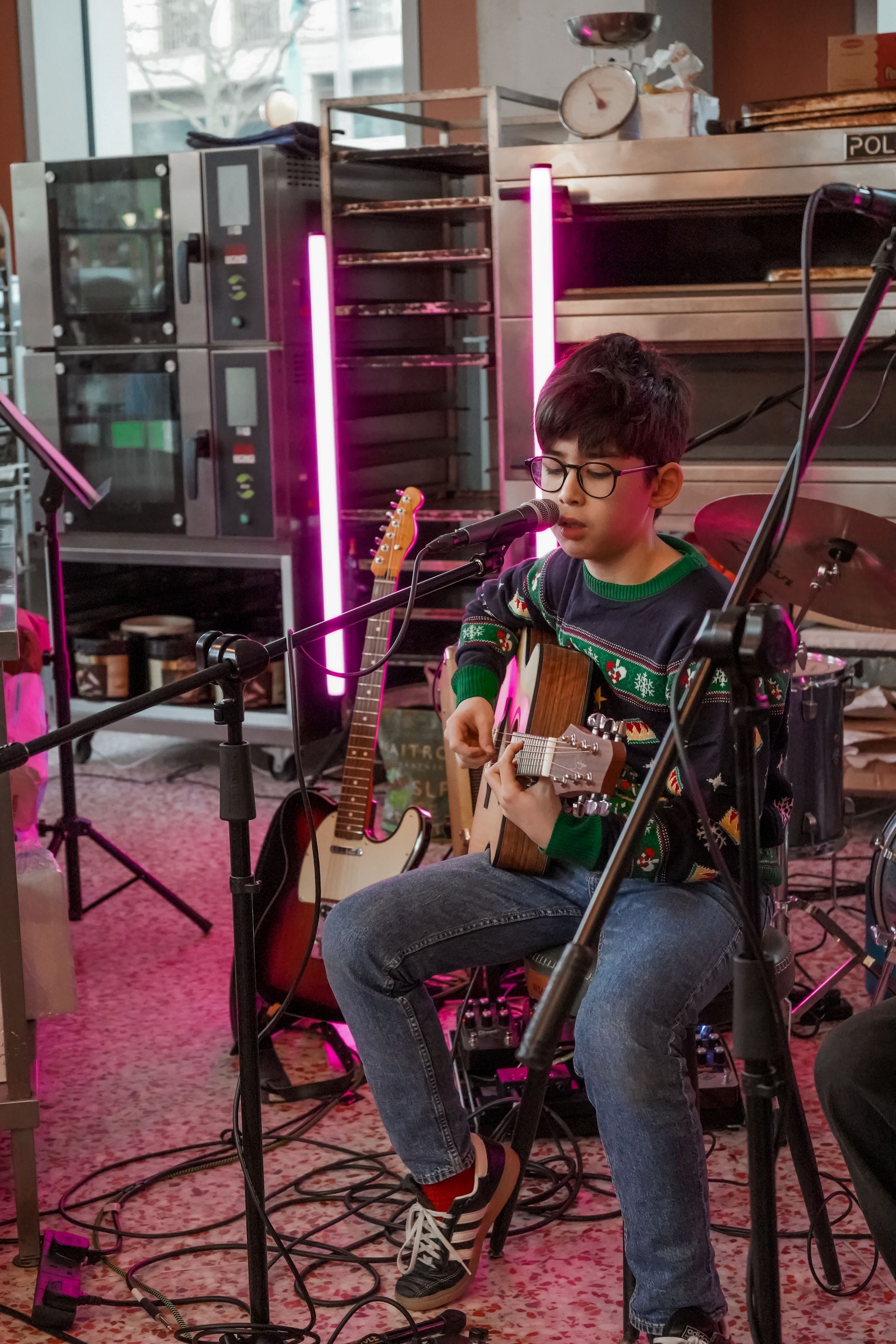 A person in a holiday sweater plays an acoustic guitar and sings into a microphone, surrounded by audio equipment.