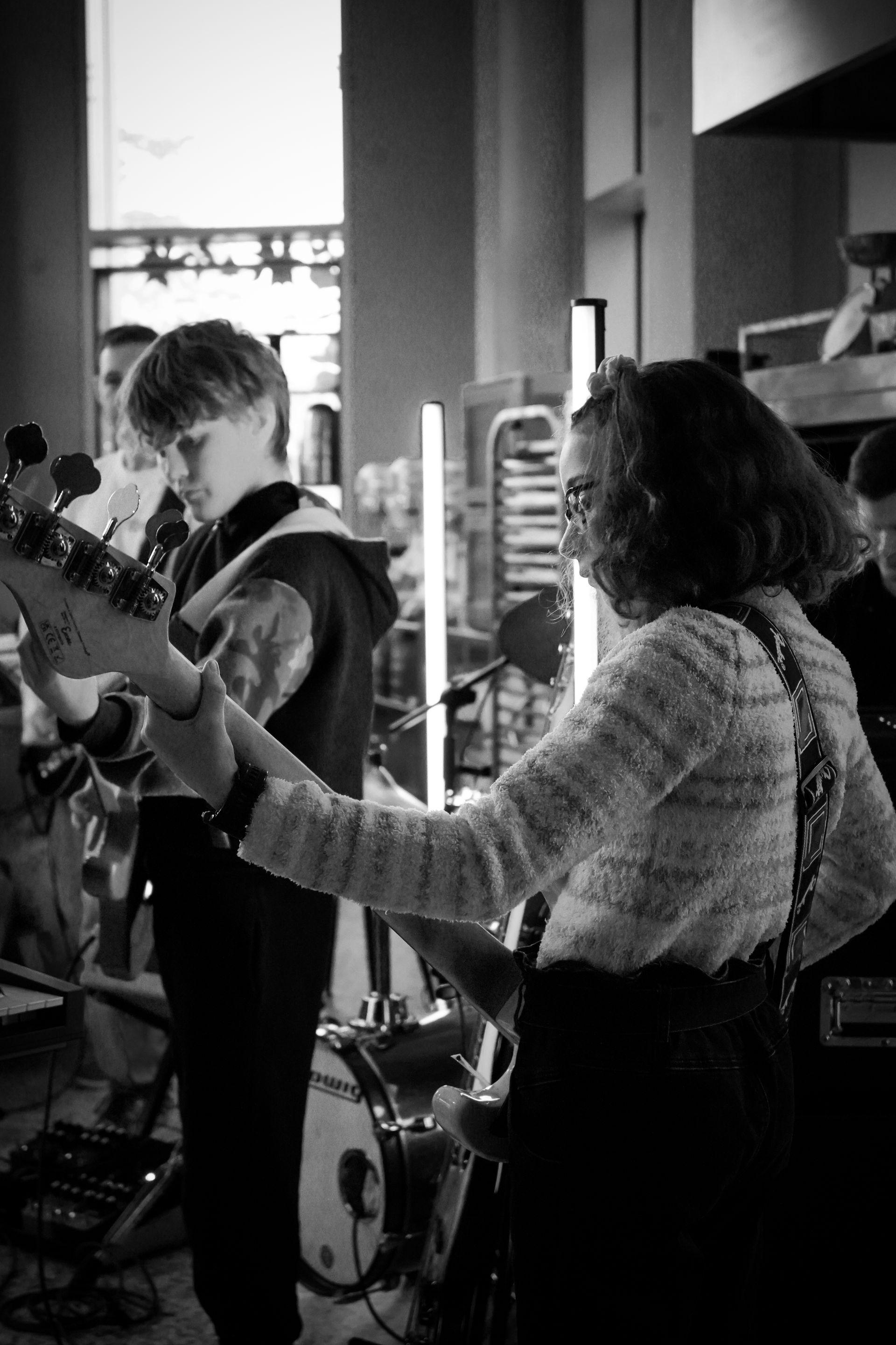 Two musicians playing guitars on a small indoor stage, with drums and speakers behind them.