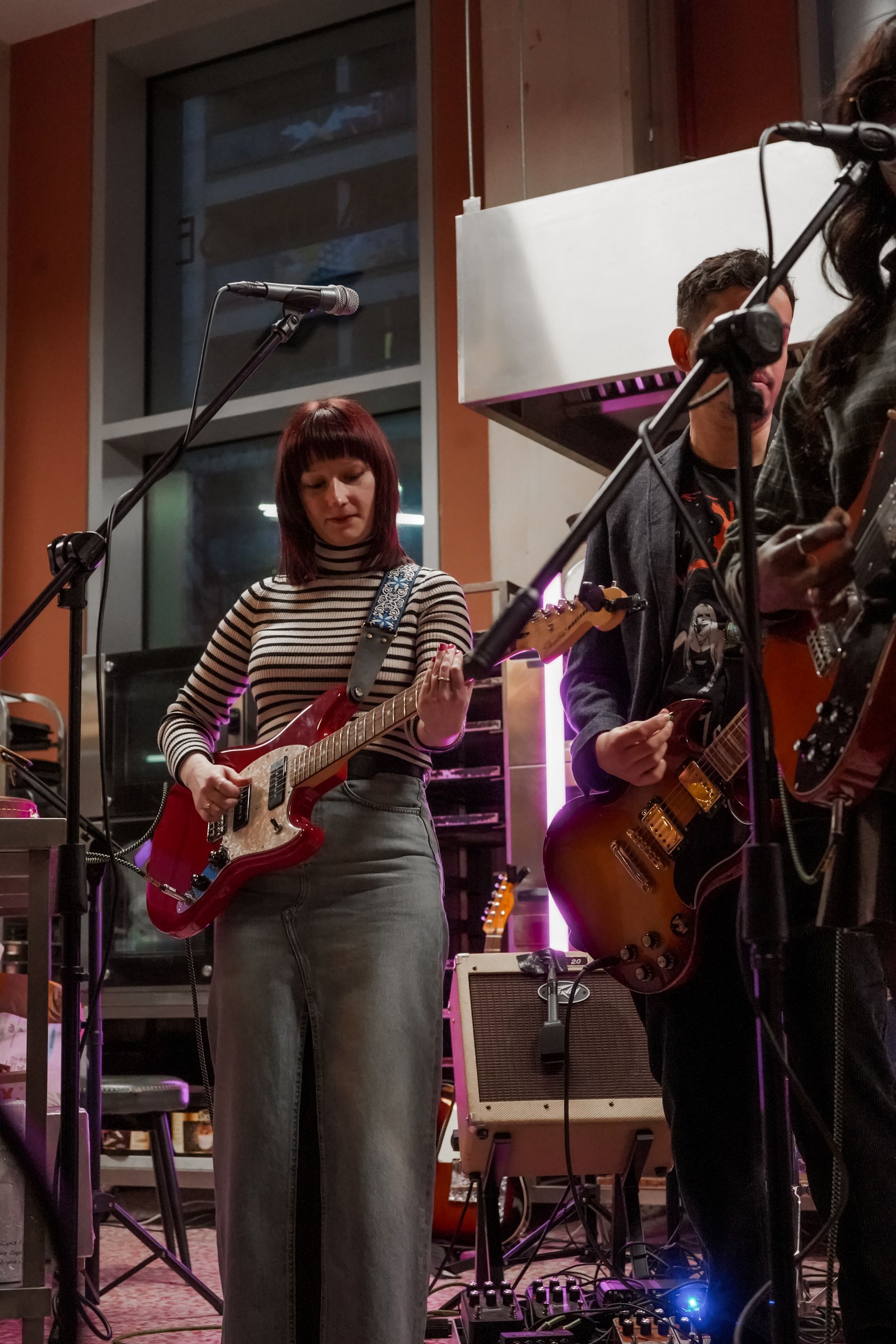 Musicians playing guitars on a small indoor stage, with microphones and warm lighting