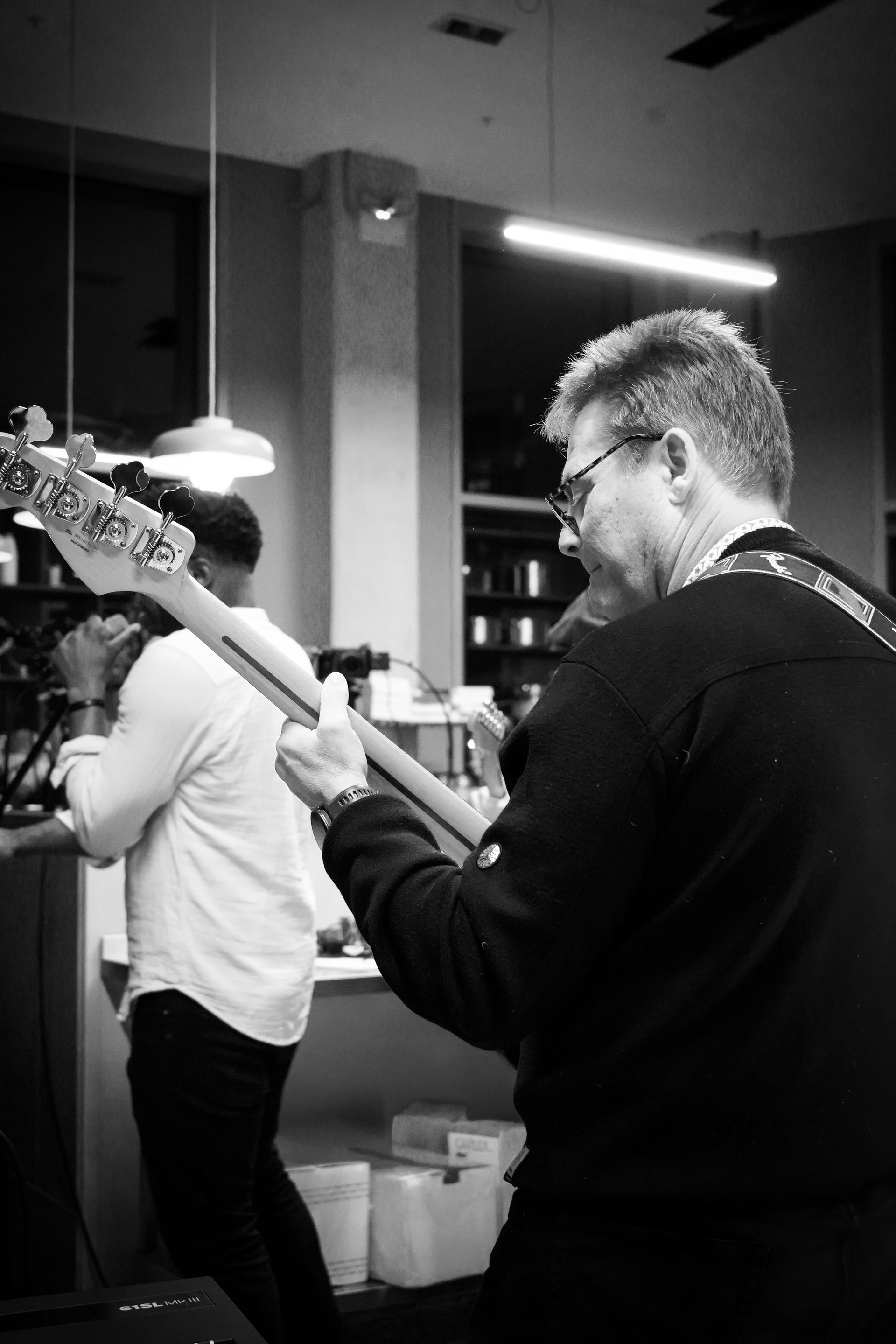Musicians playing guitar in a dim indoor venue, with a man in the foreground and another behind him.