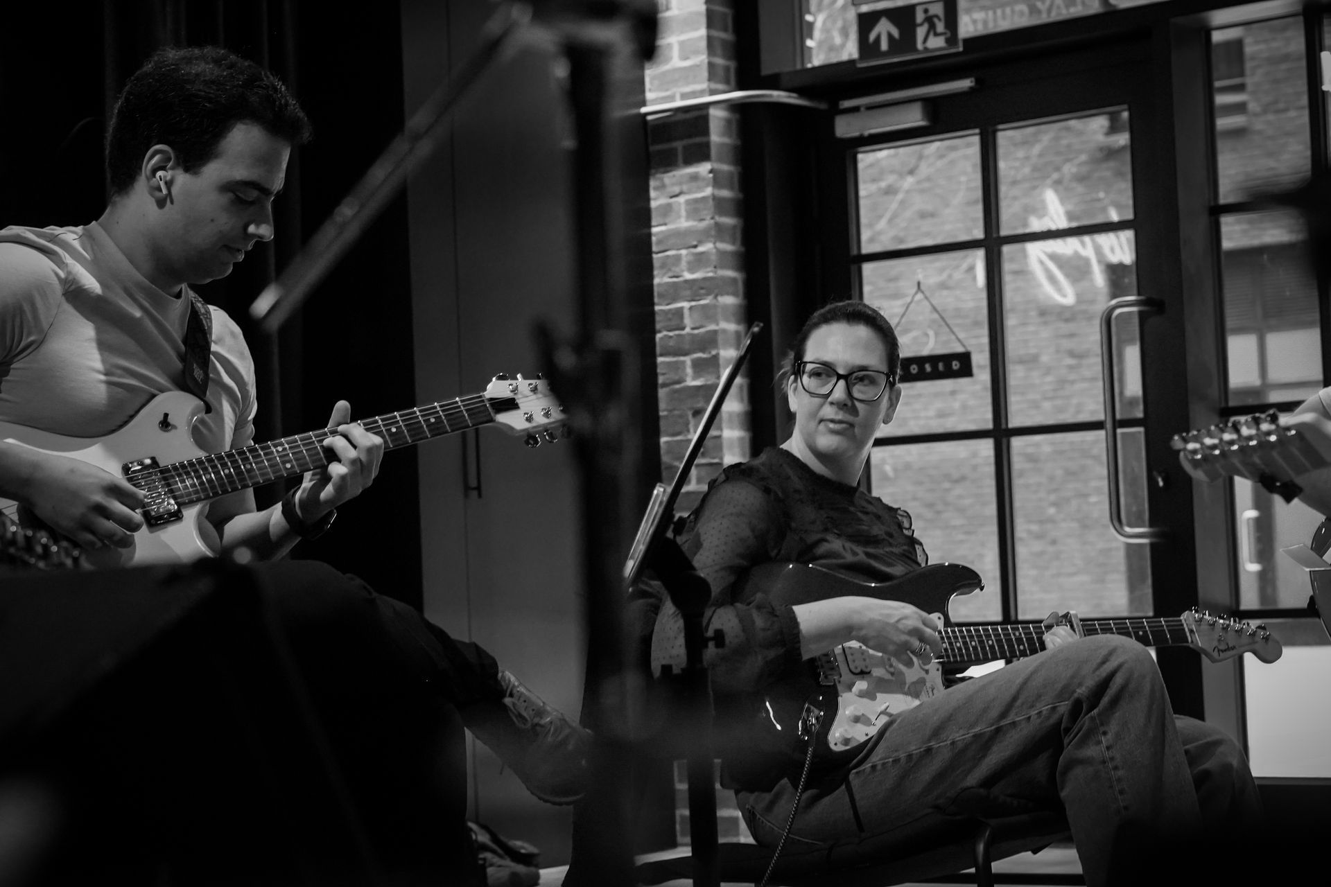 Two musicians playing electric guitars in a rehearsal space, with one looking towards the other.