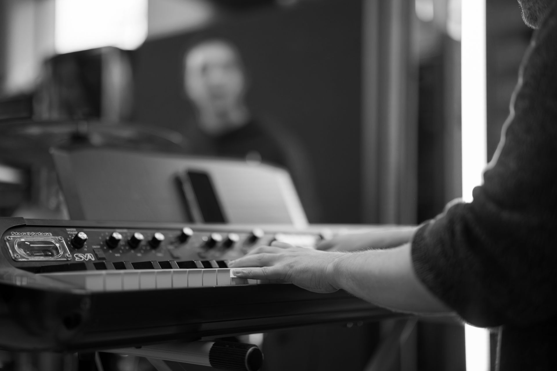 Person playing a synthesizer keyboard, with blurred reflection of another person in the background.