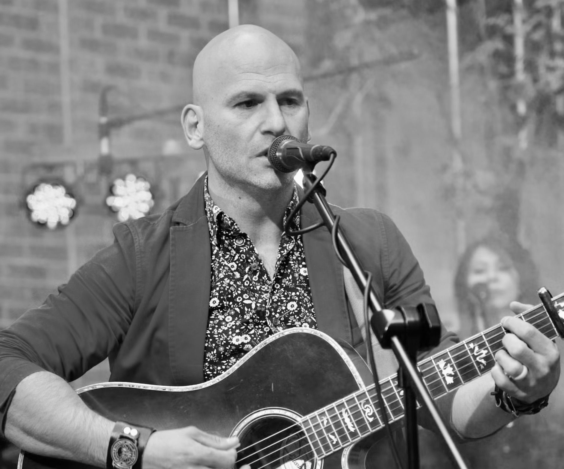 Bald man singing and playing acoustic guitar on stage, wearing a patterned shirt and blazer.