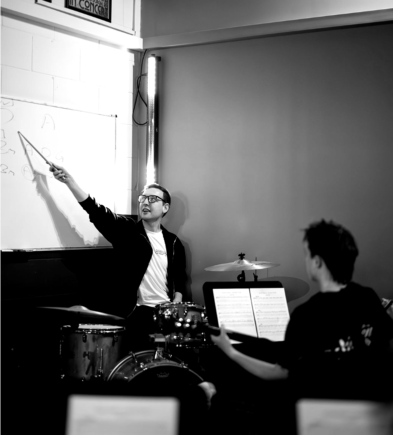 Drum teacher pointing at whiteboard, instructing student during a lesson, drum set visible.