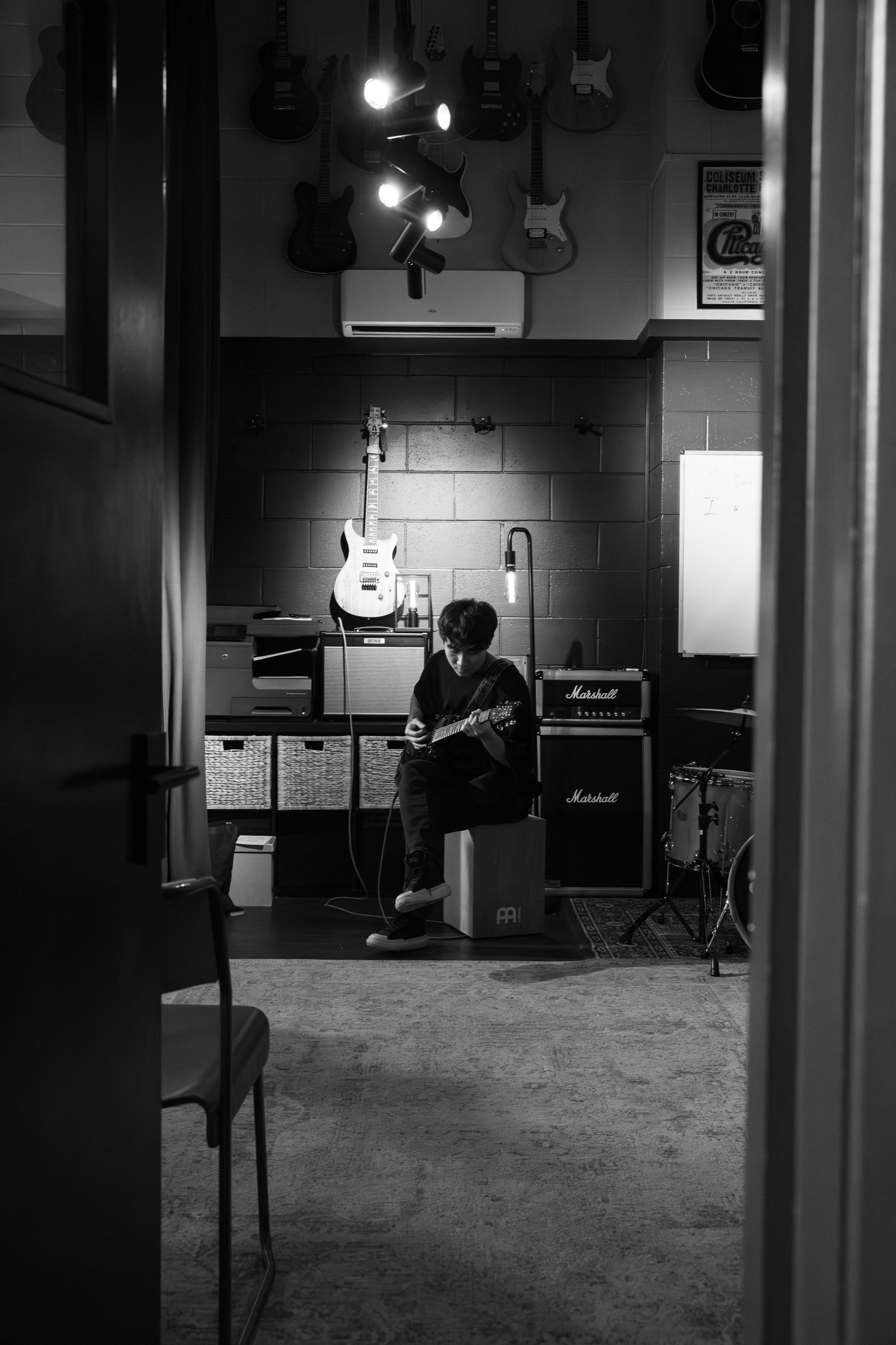 A musician sits in a recording studio, playing an instrument. Guitars hang on the wall, Marshall amps nearby.