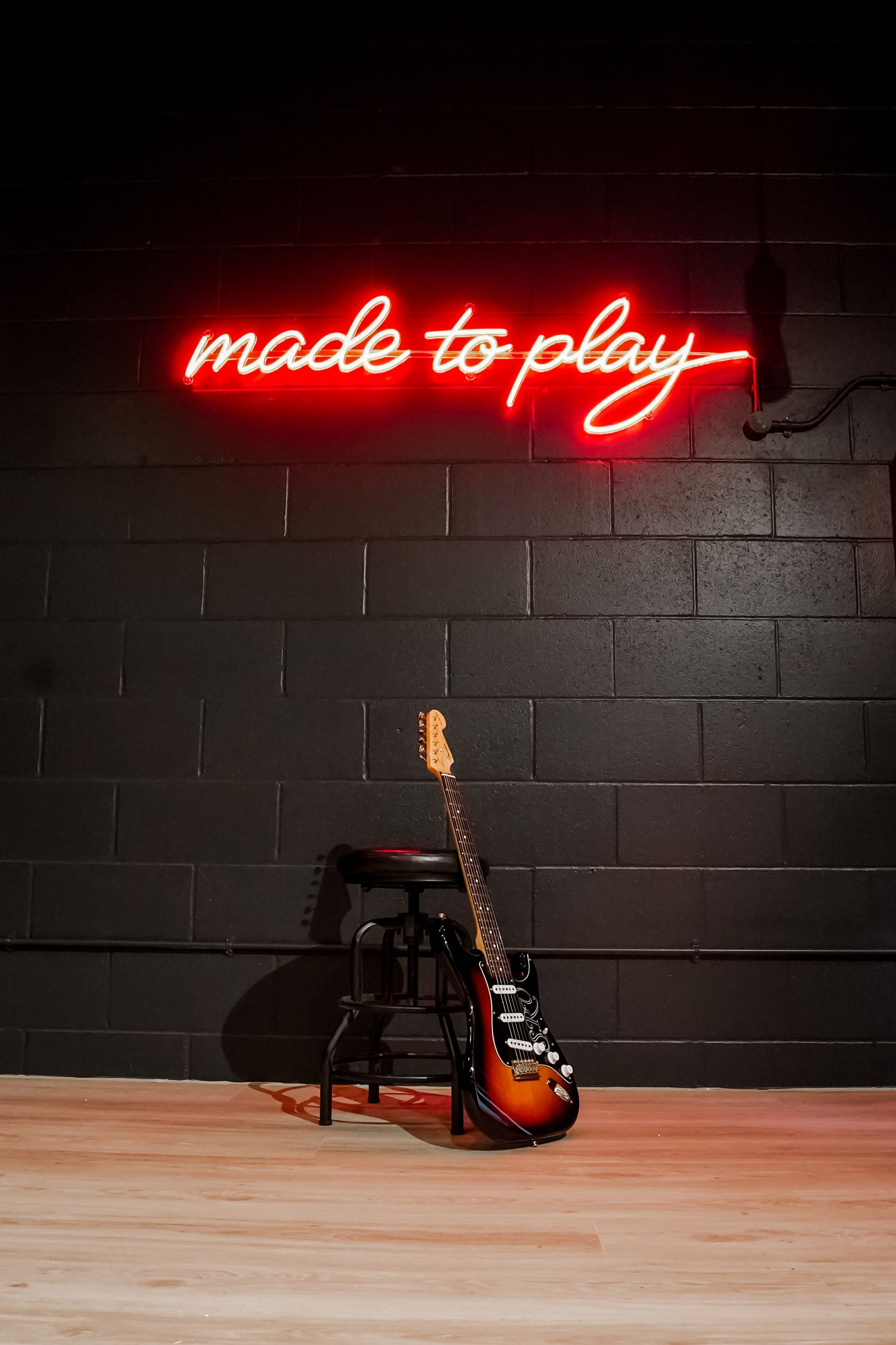 Neon “made to play” sign above an electric guitar and stool on a dark stage-like wall