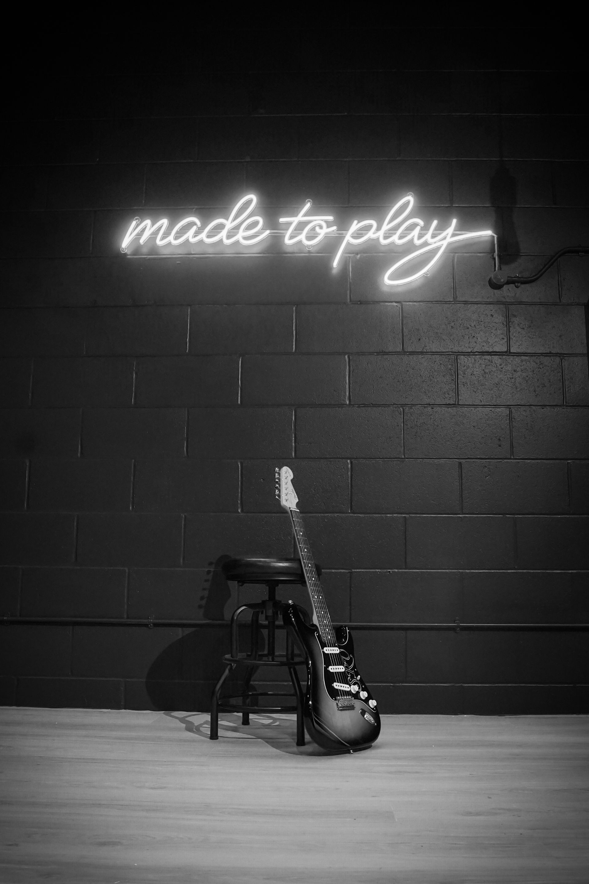 An electric guitar leans against a small stool in front of a dark brick wall featuring a neon