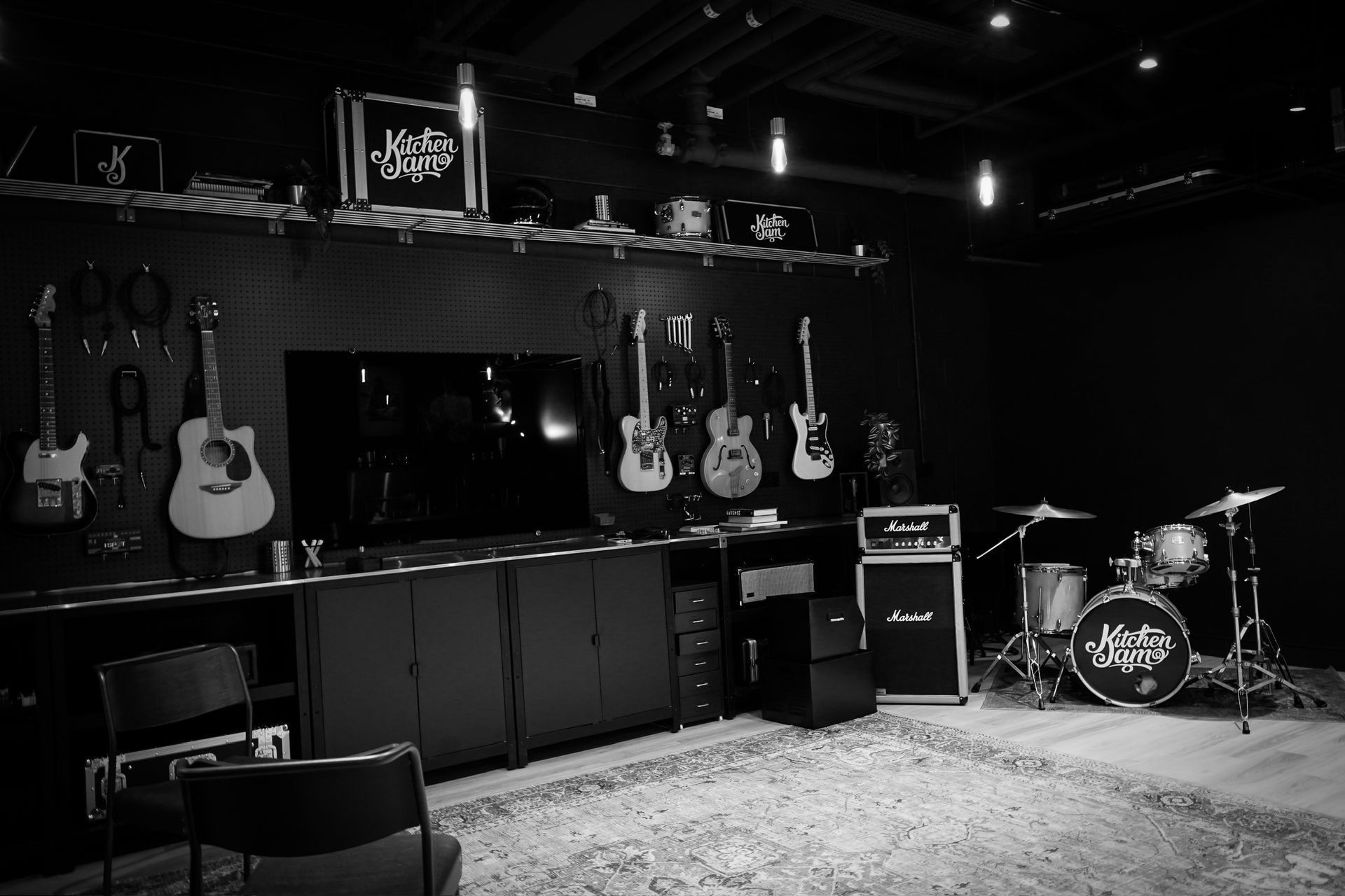Black and white wide shot of a music studio with guitar, bass, amps and a drum kit.