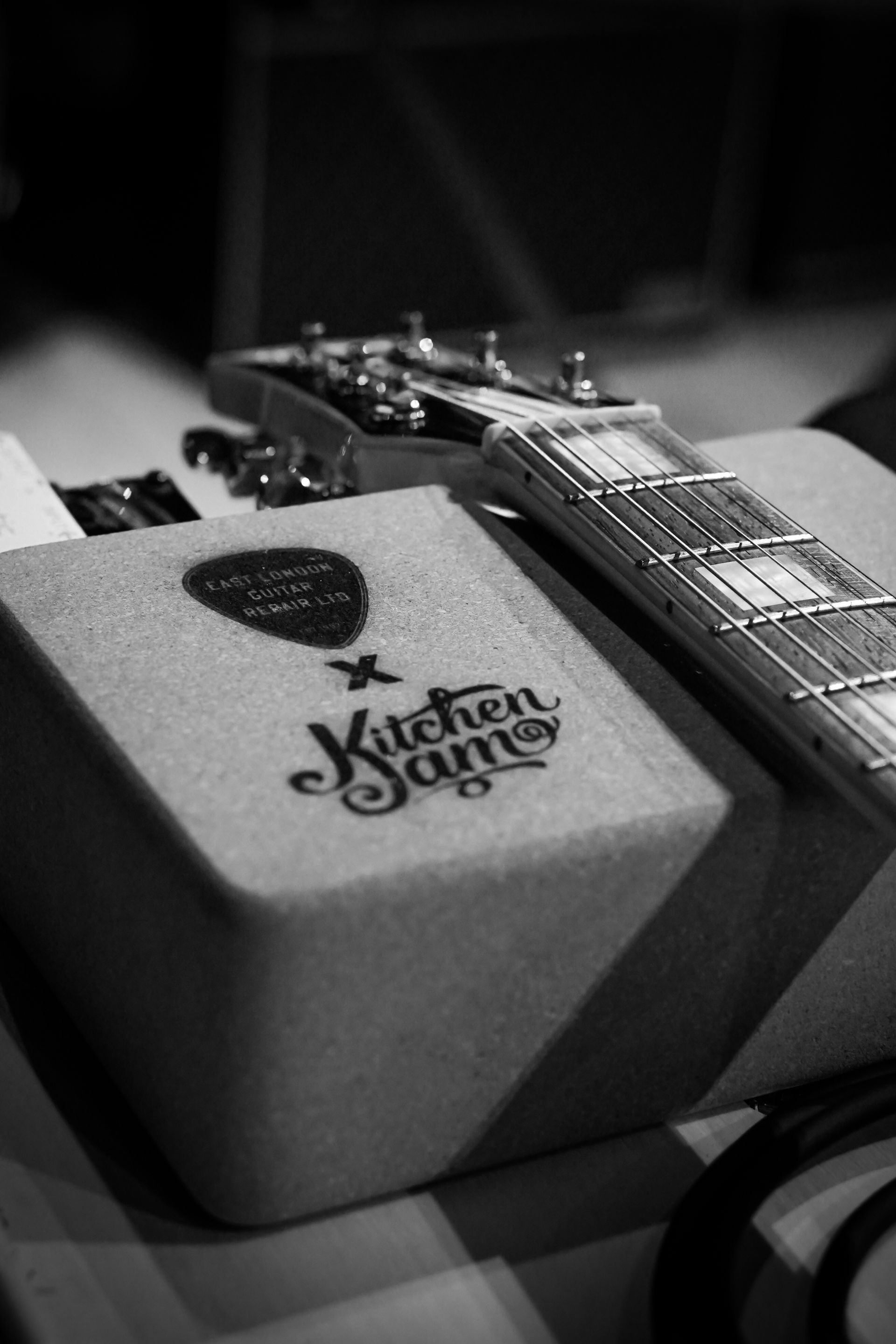 Black-and-white close-up of a guitar neck resting on a foam block with a logo