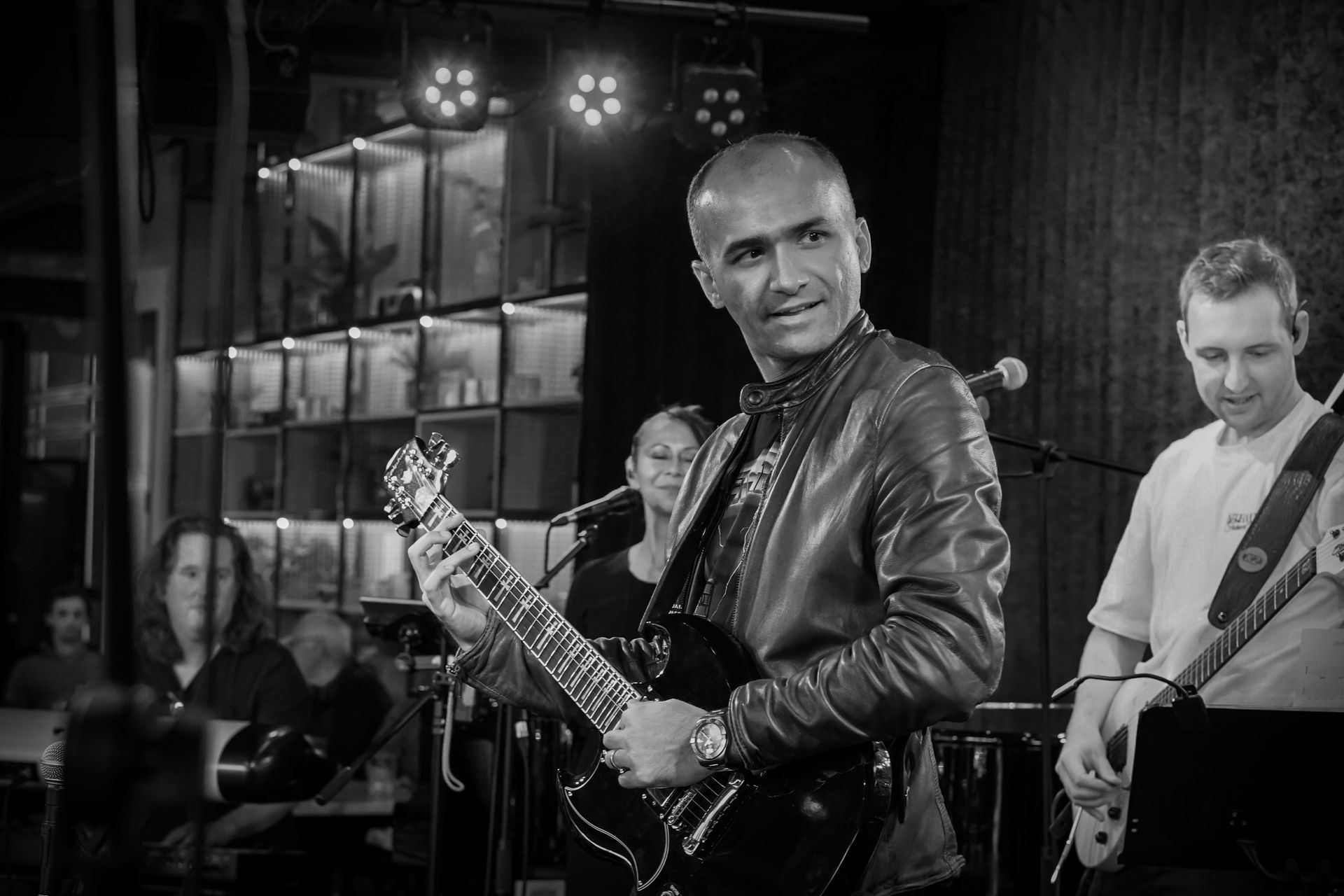 Musician holding a guitar smiles onstage during a live performance, with band members and lights behind him