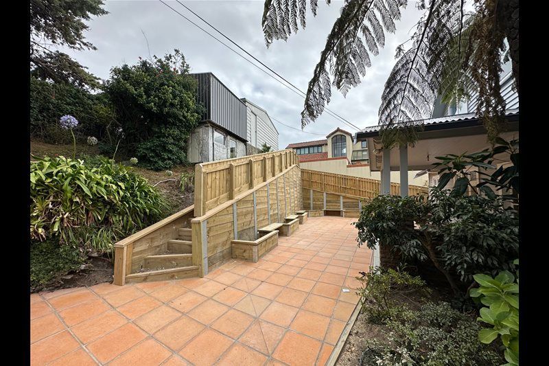 Terracotta-tiled outdoor ramp with wooden handrails beside a building and greenery