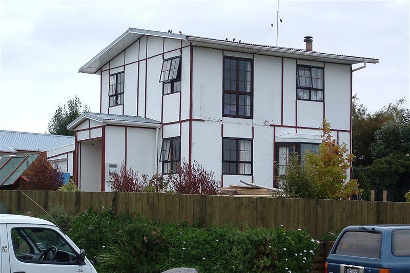 Two-story white house with red trim behind a wooden fence, with cars parked in front