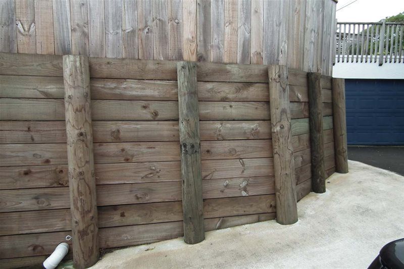 Wooden retaining wall with thick round posts beside a paved area and blue gate.