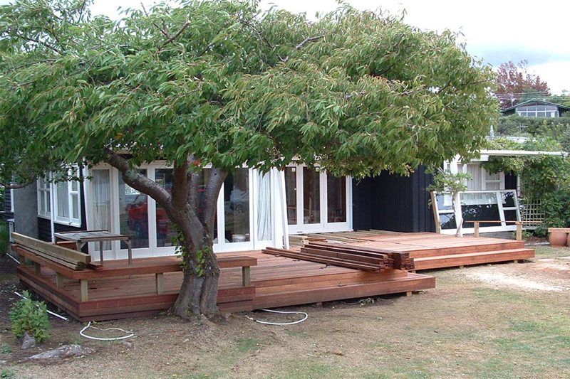 House with large tree over wooden deck and patio seating in a yard
