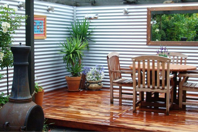 Wooden deck with outdoor dining table, chairs, potted plants, and corrugated metal walls under a covered patio