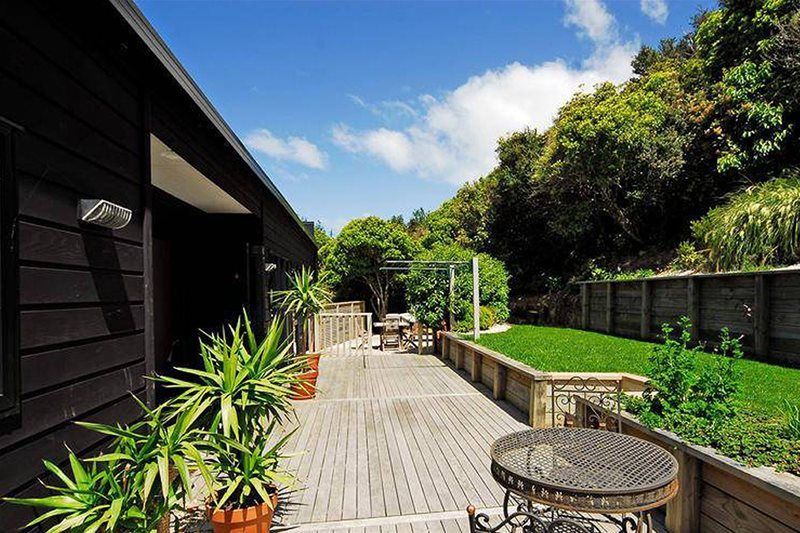 Wooden patio beside a dark house, with potted plants, railing, and a green garden under a blue sky.