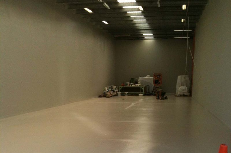 Empty warehouse interior with concrete walls, bright overhead lights, and equipment stacked at the far end.