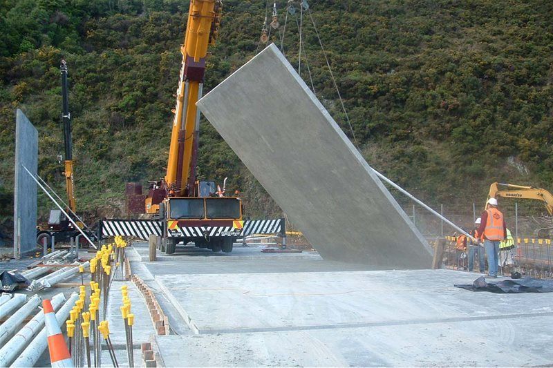 Construction workers use a crane to lift a large concrete panel on a road site.