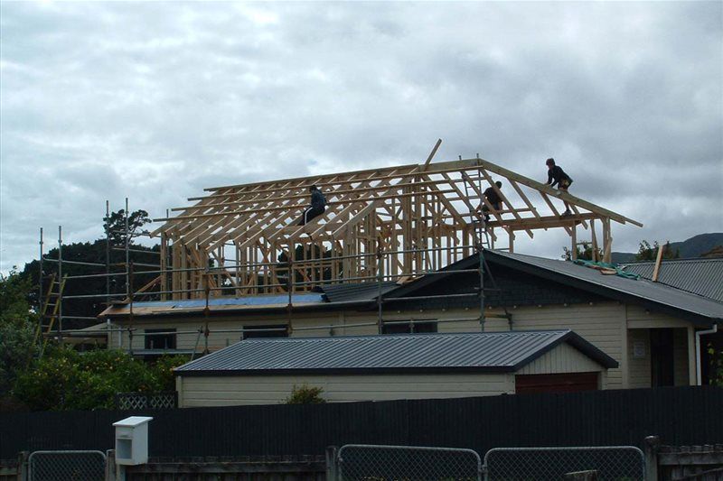 House under construction with exposed wooden roof framing above a dark-roofed home and cloudy sky.