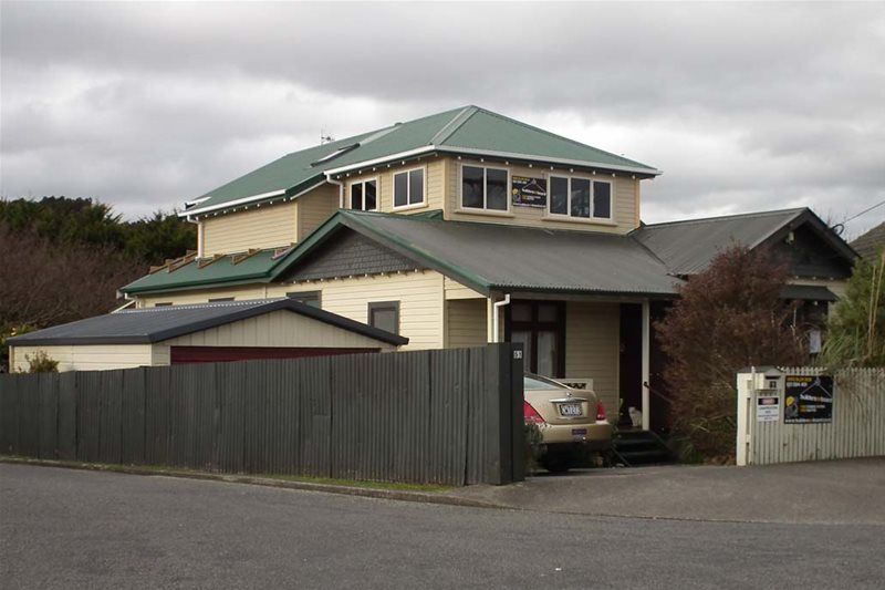 Two-story beige house with green roof behind a gray fence on a cloudy day