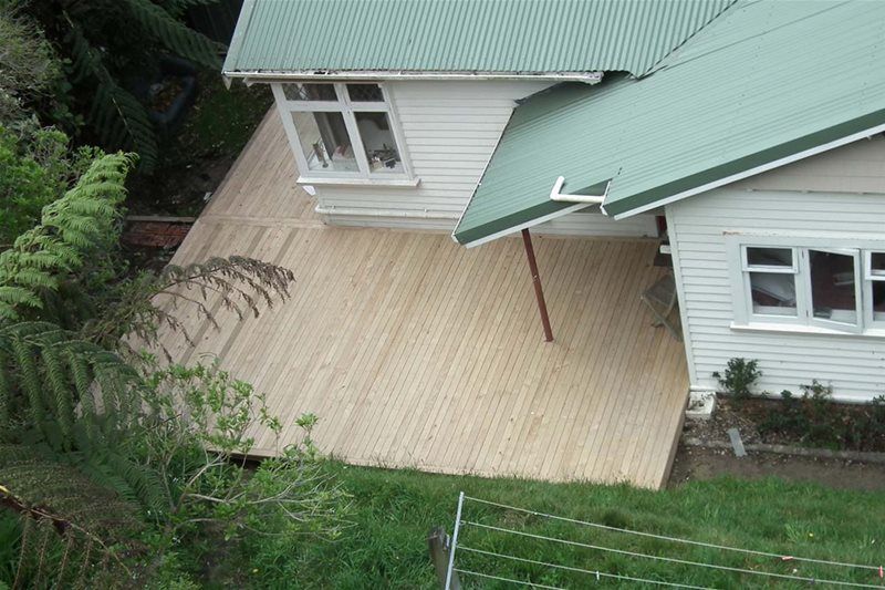 Backyard deck with tan wooden floor beside a white house with green roof, viewed from above