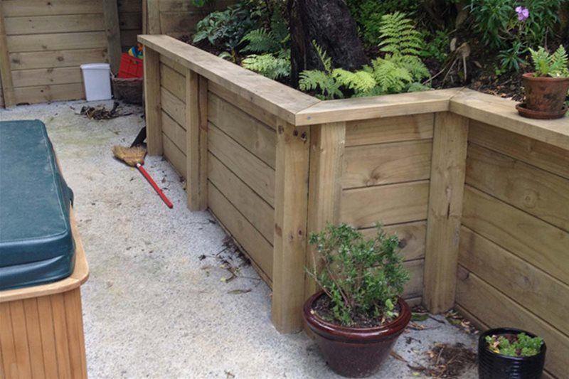 Wooden garden retaining wall beside a patio, with potted plants and a covered pool on the left.