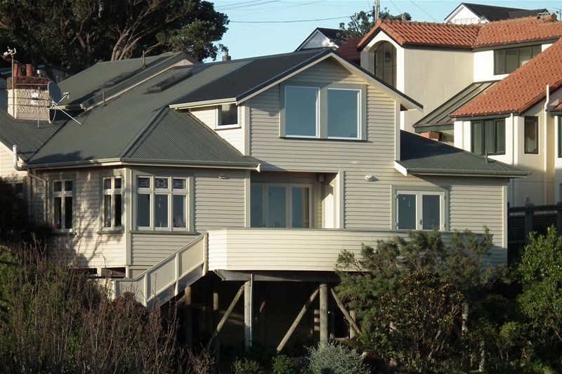Two-story beige house with a dark roof and white trim on a hillside, surrounded by trees and neighboring homes